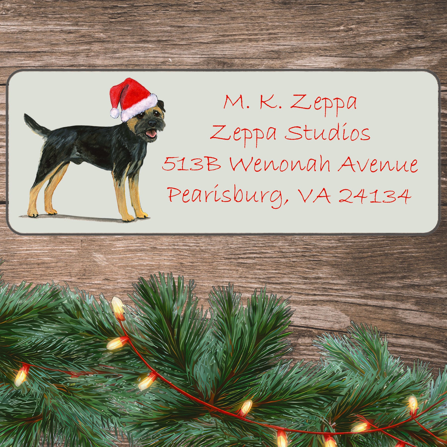 An illustration of a black and tan Border Collie wearing a Santa Hat on an ivory return address label set off by a wood surface with holiday greenery background.