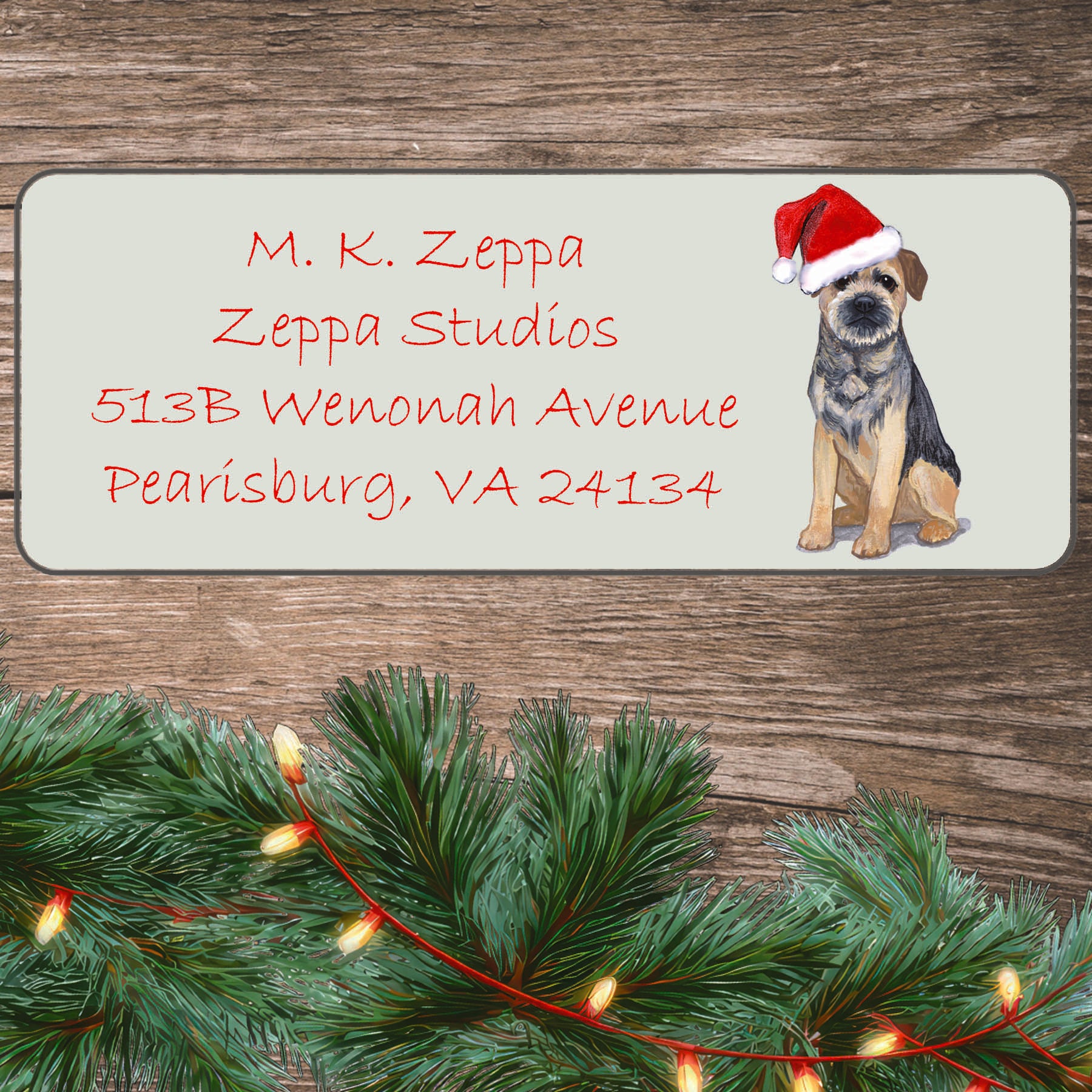 An illustration of a Border Terrier wearing a Santa Hat on an ivory return address label set off by a wood surface with holiday greenery background.
