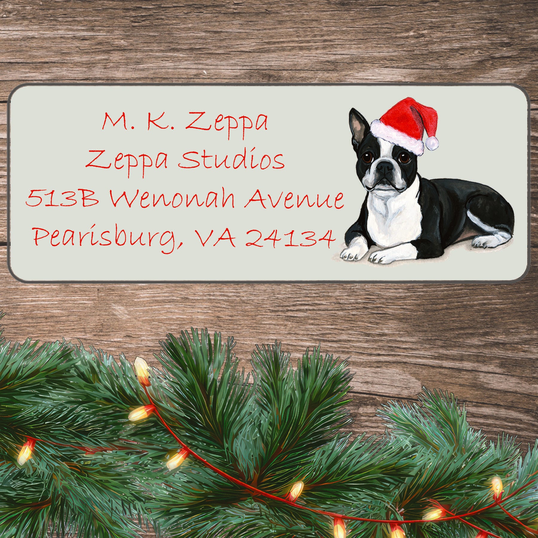 An illustration of a Boston Terrier lying down wearing a Santa Hat on an ivory return address label set off by a wood surface with holiday greenery background.