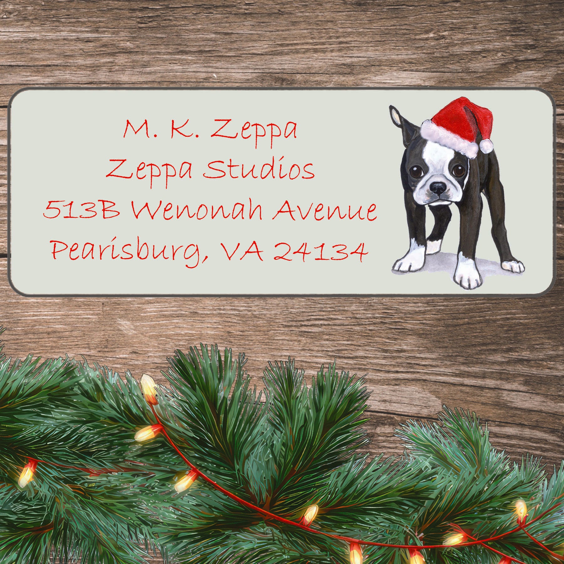 An illustration of a Boston Terrier puppy wearing a Santa Hat on an ivory return address label set off by a wood surface with holiday greenery background.