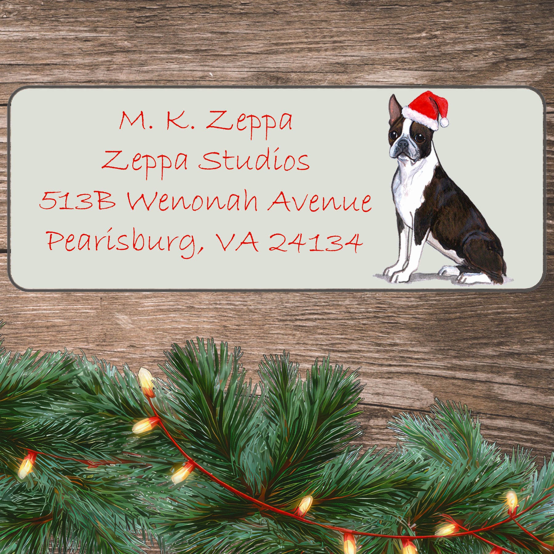 An illustration of a Boston Terrier wearing a Santa Hat on an ivory return address label set off by a wood surface with holiday greenery background.