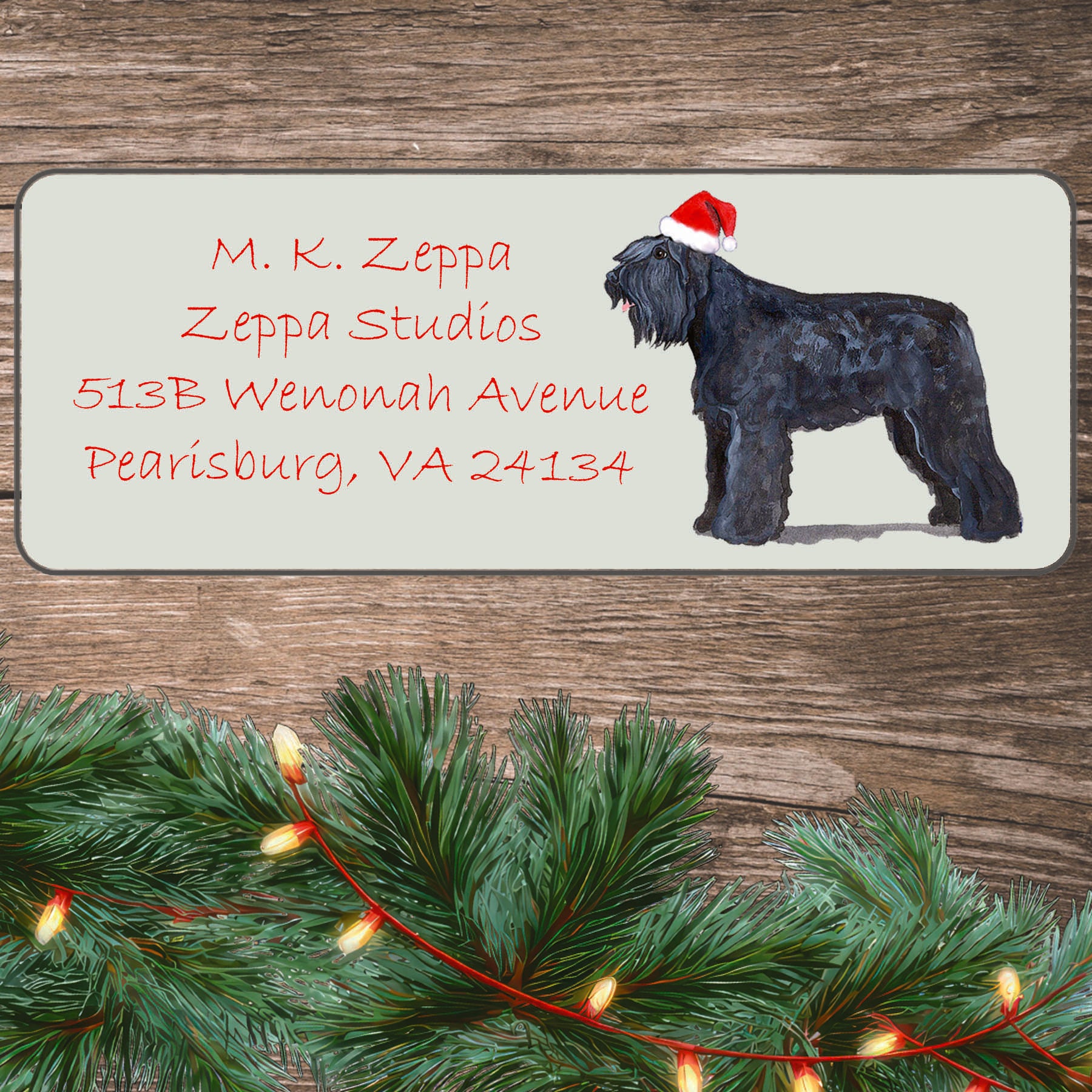 An illustration of a Bouvier wearing a Santa Hat on an ivory return address label set off by a wood surface with holiday greenery background.