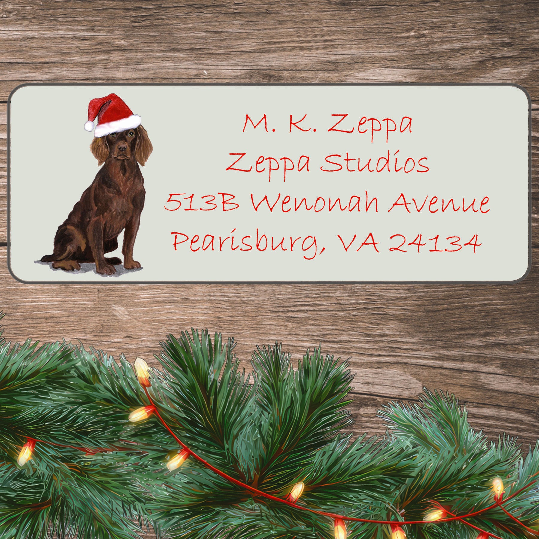 An illustration of a Boykin Spaniel wearing a Santa Hat on an ivory return address label set off by a wood surface with holiday greenery background.