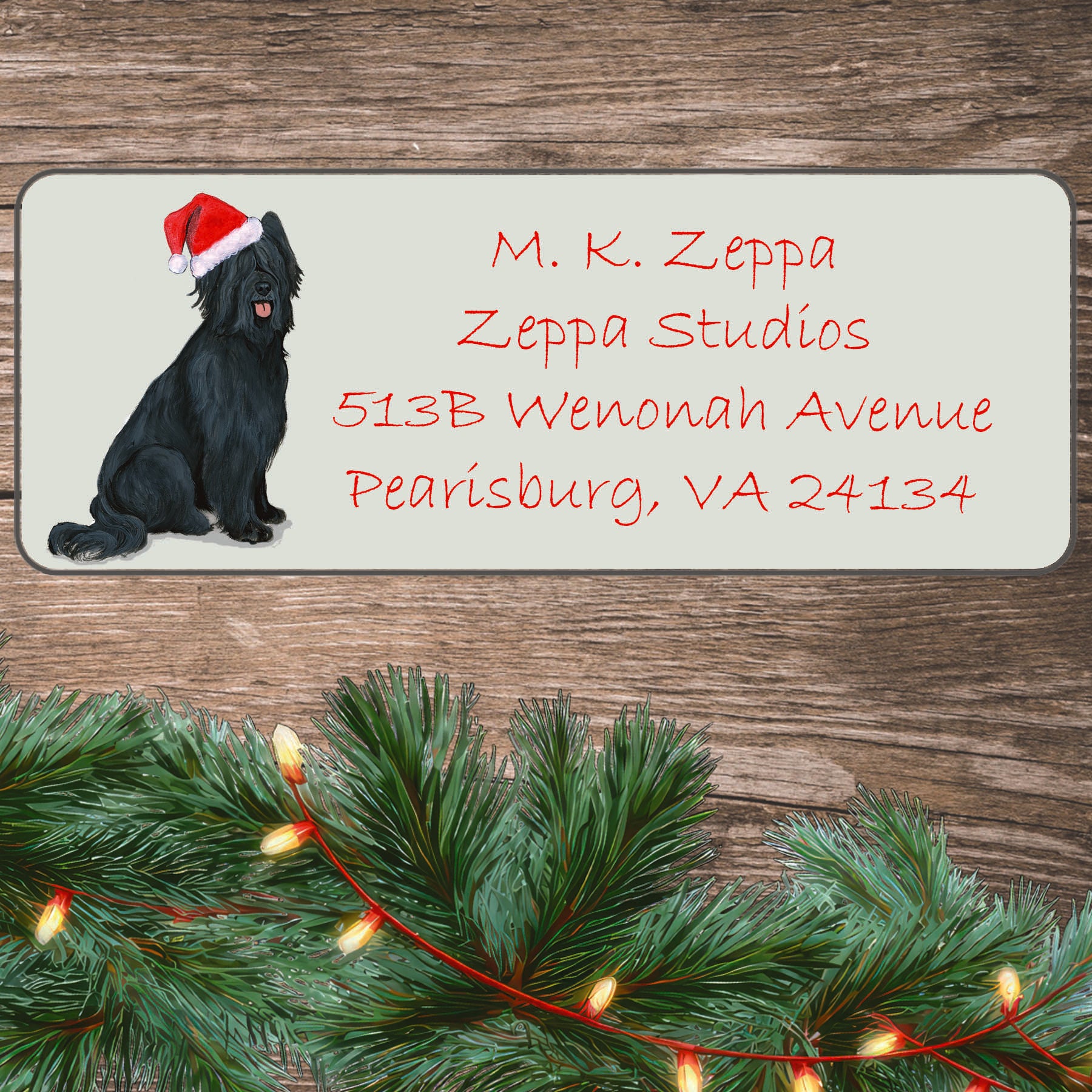 An illustration of a black Briard wearing a Santa Hat on an ivory return address label set off by a wood surface with holiday greenery background.