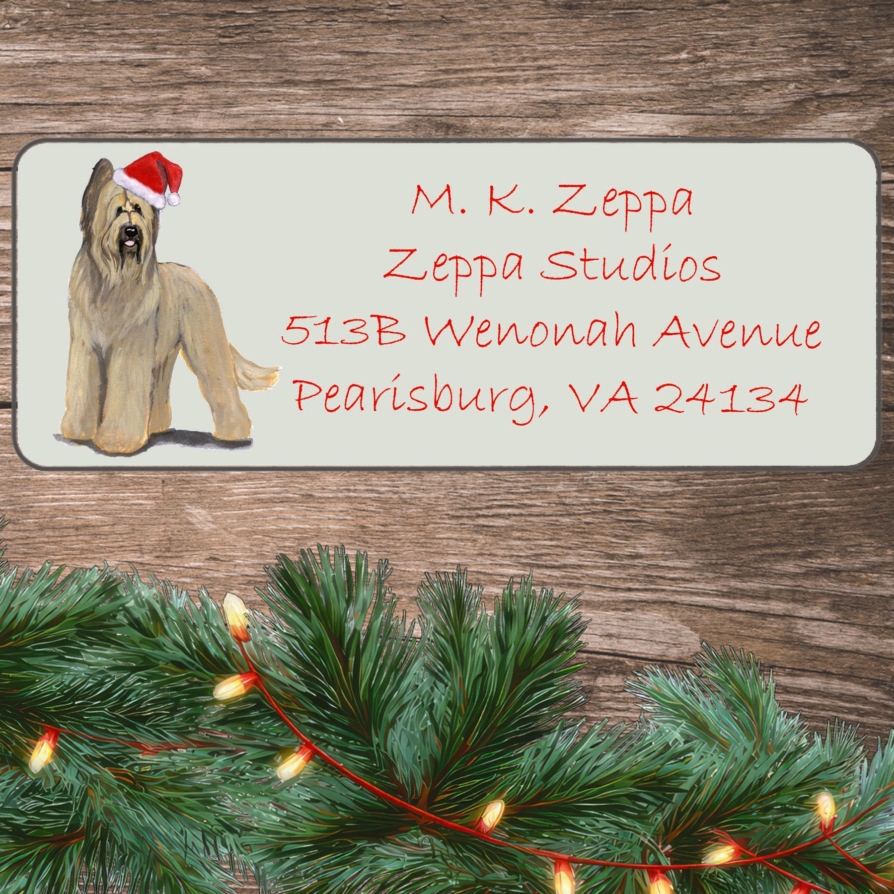 An illustration of a Briard wearing a Santa Hat on an ivory return address label set off by a wood surface with holiday greenery background.
