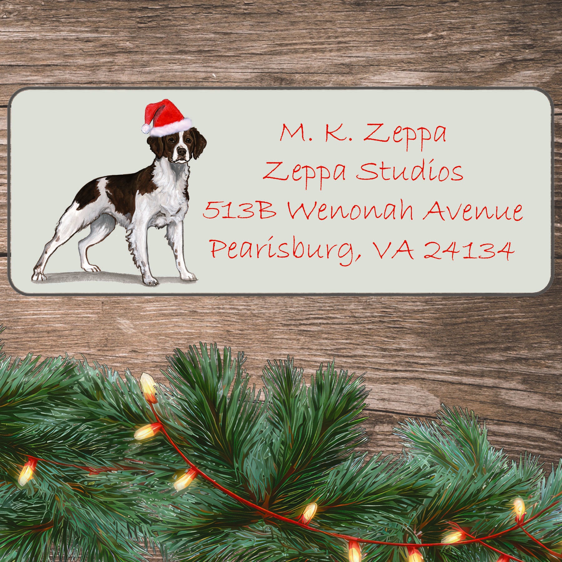 An illustration of a liver and white Brittany wearing a Santa Hat on an ivory return address label set off by a wood surface with holiday greenery background.