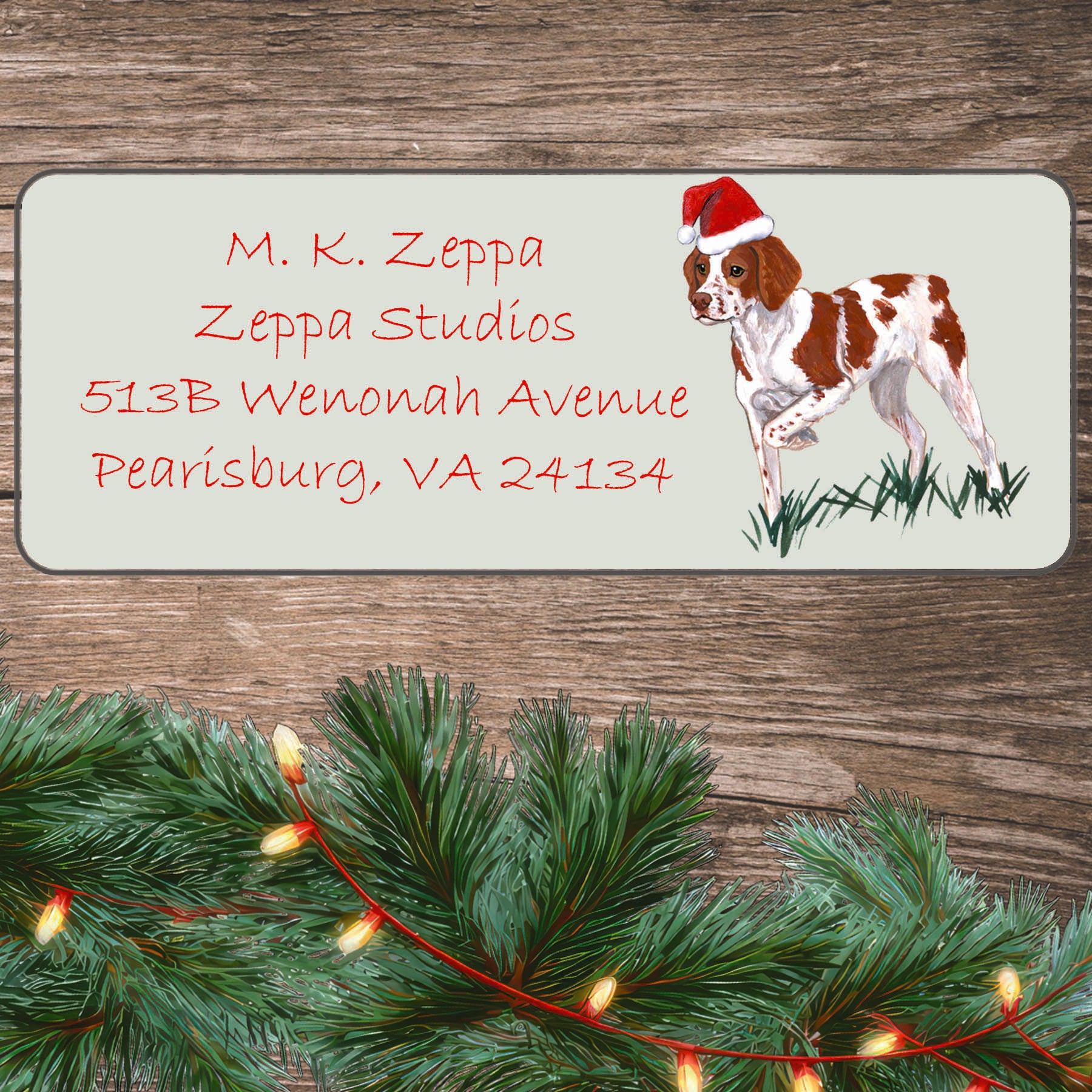 An illustration of a Brittany wearing a Santa Hat on an ivory return address label set off by a wood surface with holiday greenery background.