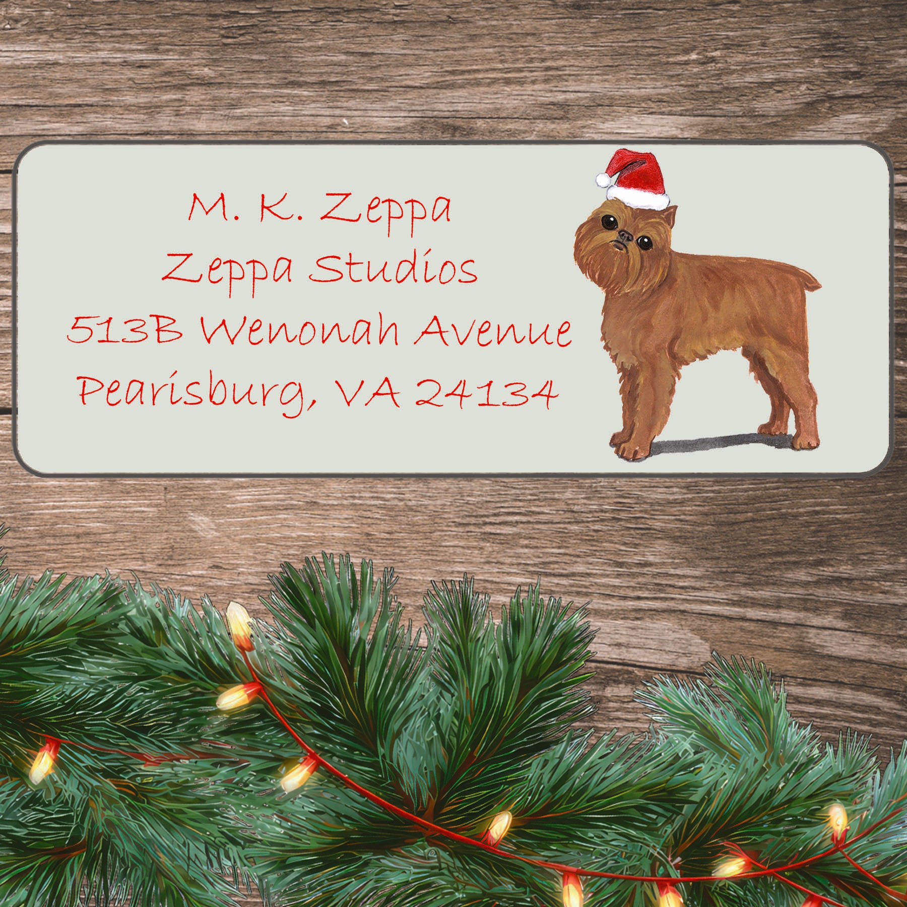 An illustration of a  Brussels Griffon wearing a Santa Hat on an ivory return address label set off by a wood surface with holiday greenery background.