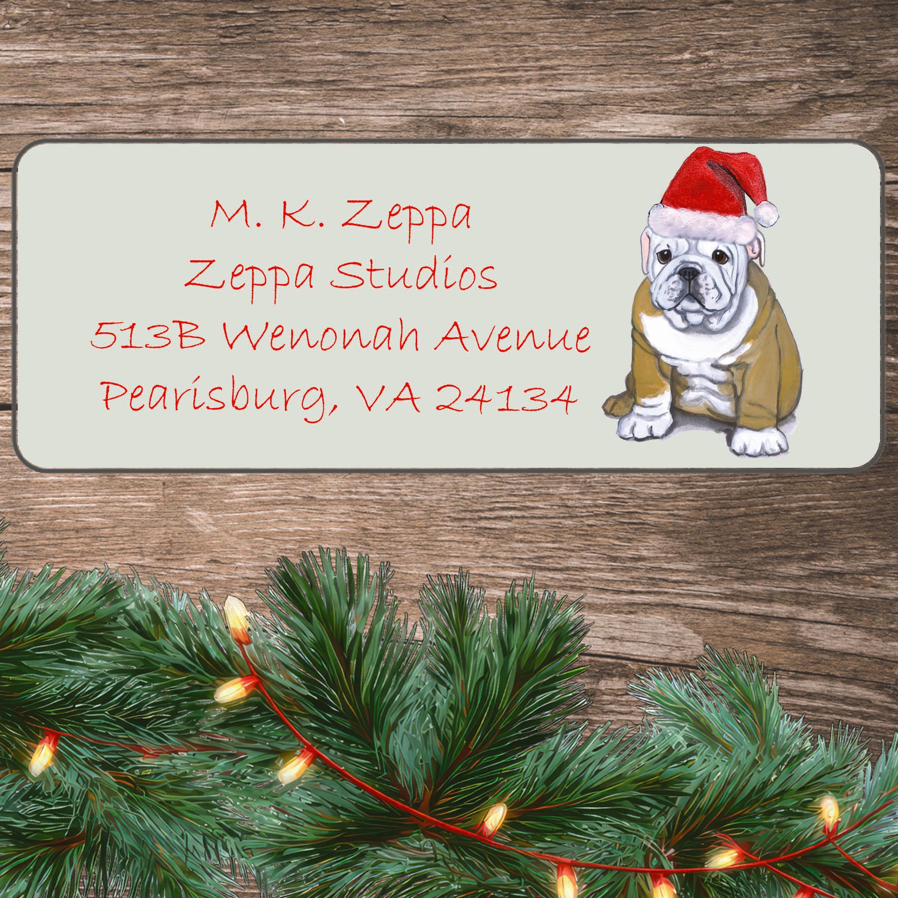 An illustration of a Bulldog puppy wearing a Santa Hat on an ivory return address label set off by a wood surface with holiday greenery background.