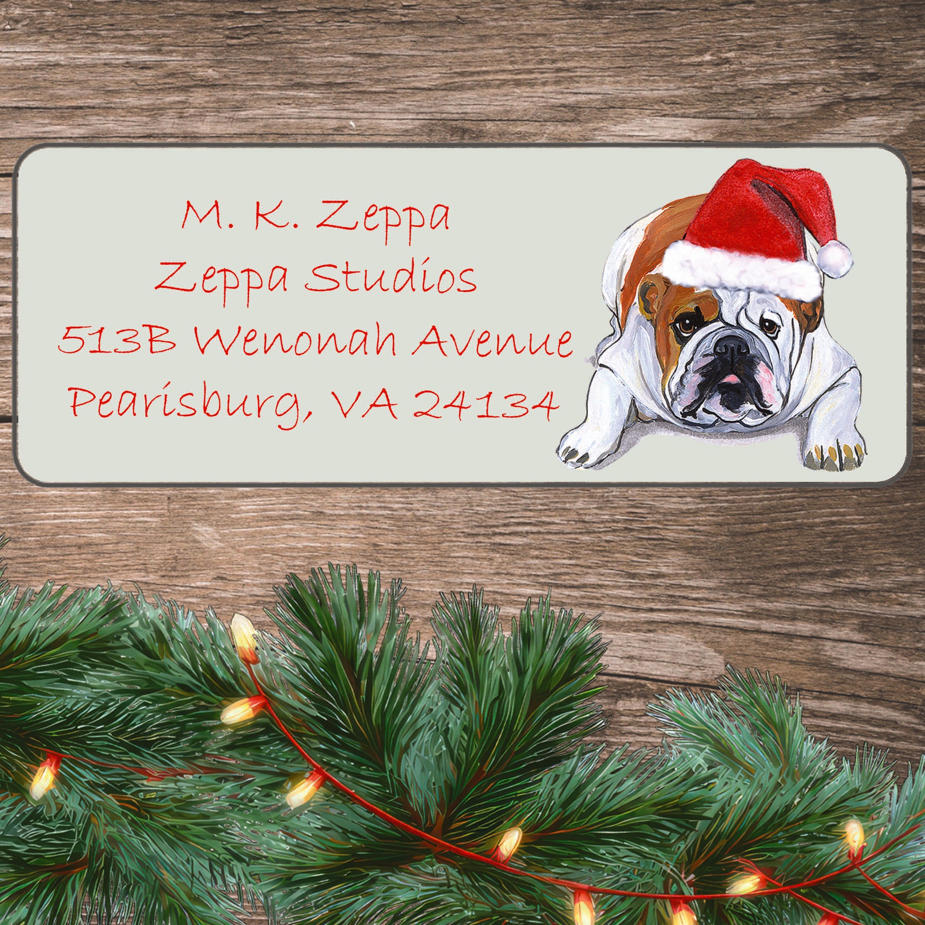 An illustration of a Bulldog wearing a Santa Hat on an ivory return address label set off by a wood surface with holiday greenery background.