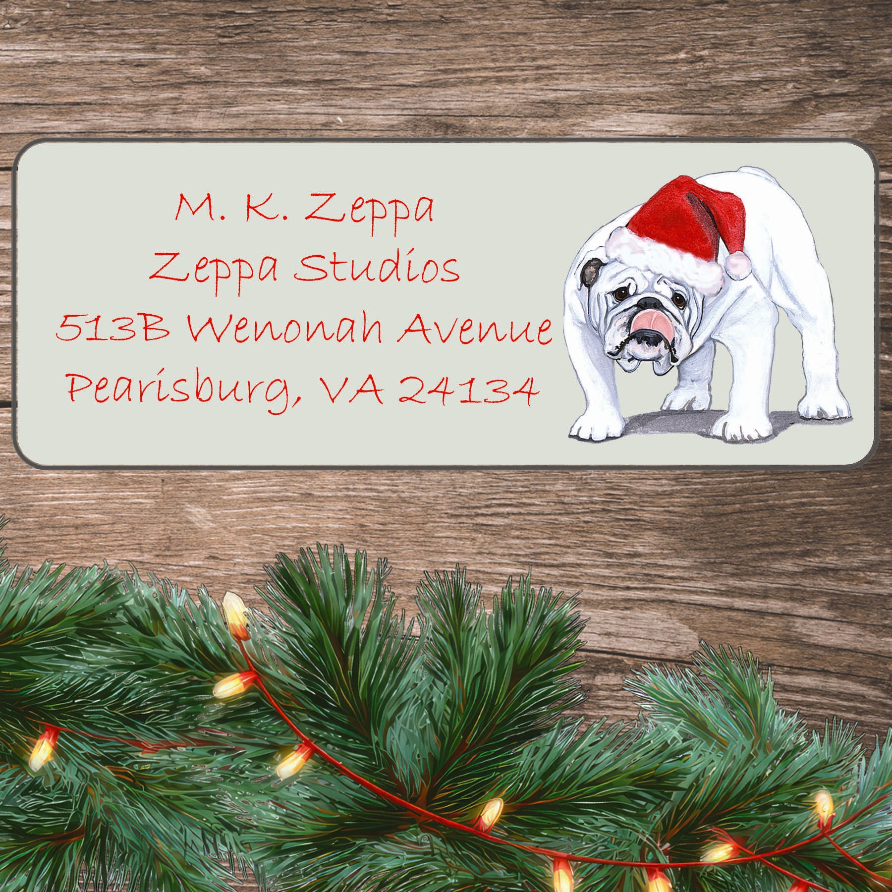An illustration of a white Bulldog wearing a Santa Hat on an ivory return address label set off by a wood surface with holiday greenery background.