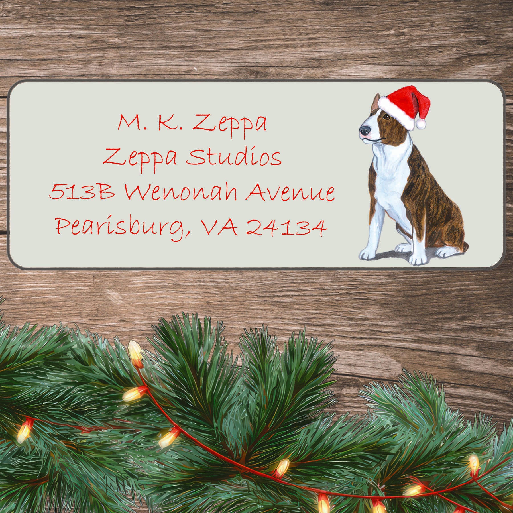 An illustration of a brindle and white Bull Terrier wearing a Santa Hat on an ivory return address label set off by a wood surface with holiday greenery background.