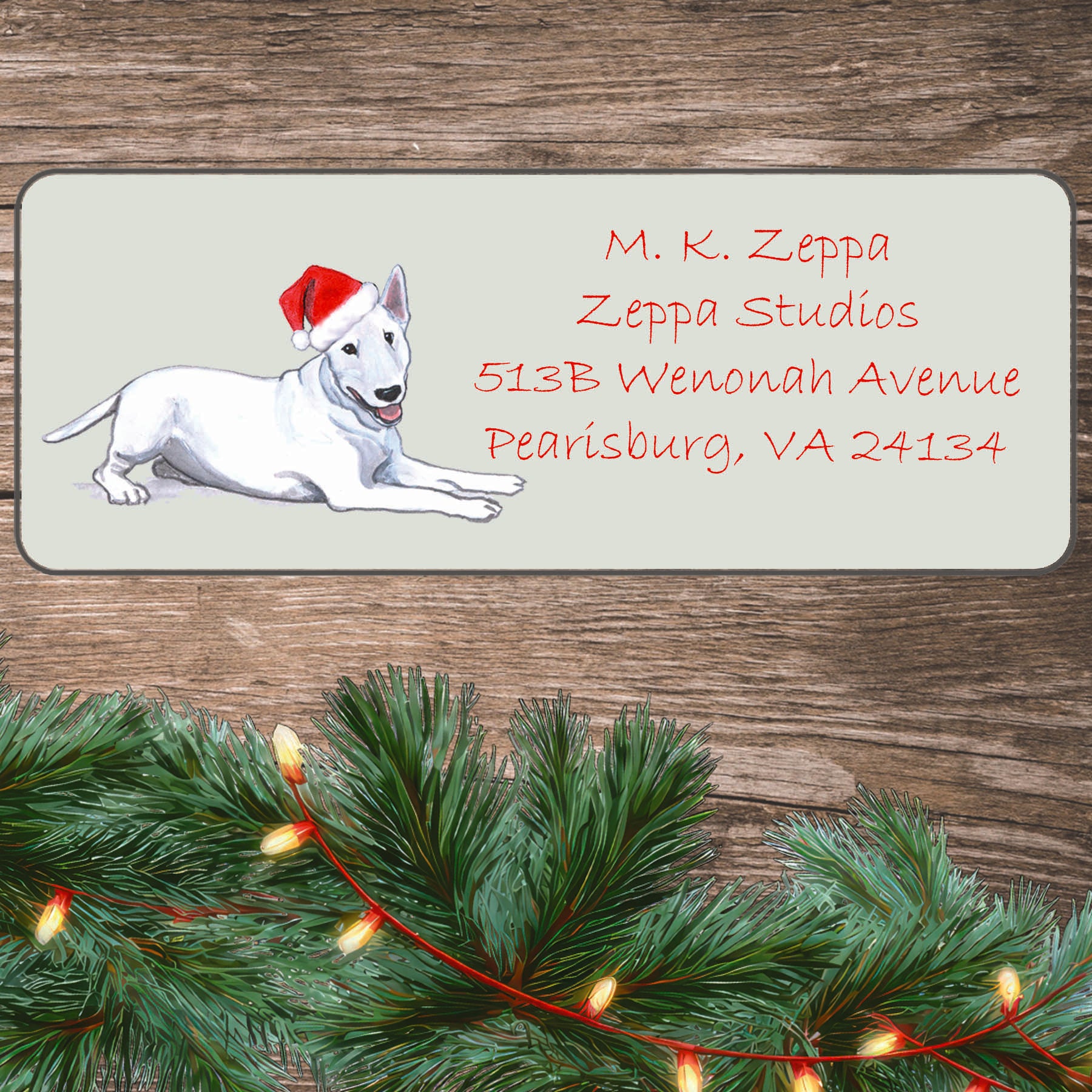 An illustration of a white Bull Terrier wearing a Santa Hat on an ivory return address label set off by a wood surface with holiday greenery background.