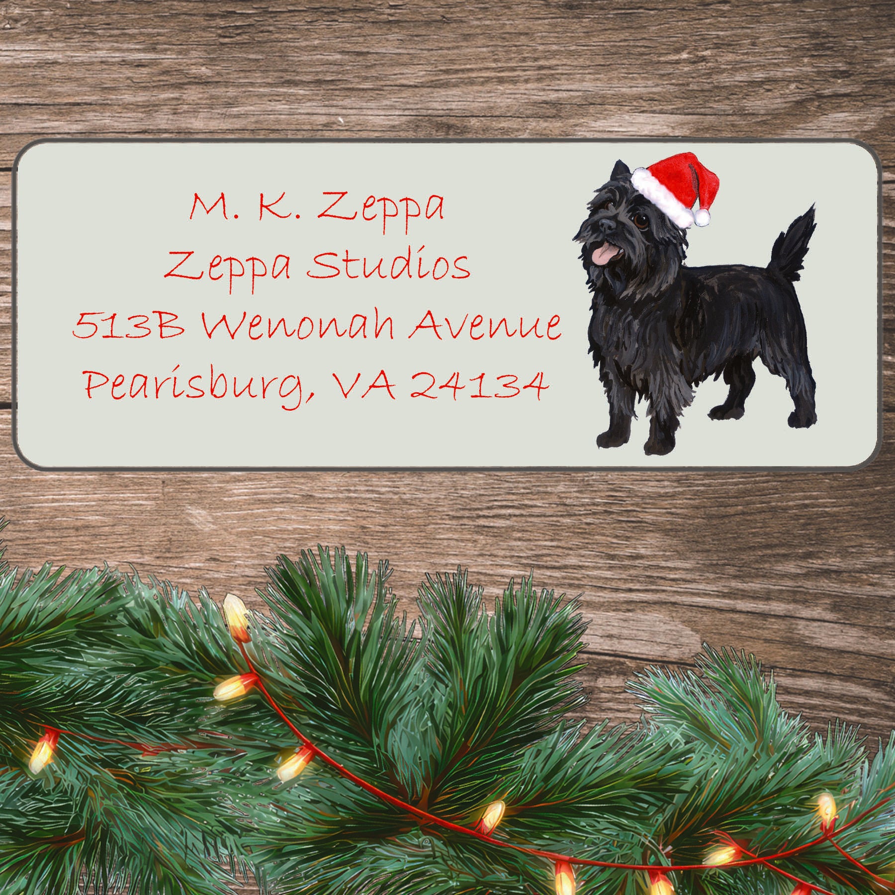 An illustration of a black Cairn Terrier wearing a Santa Hat on an ivory return address label set off by a wood surface with holiday greenery background.