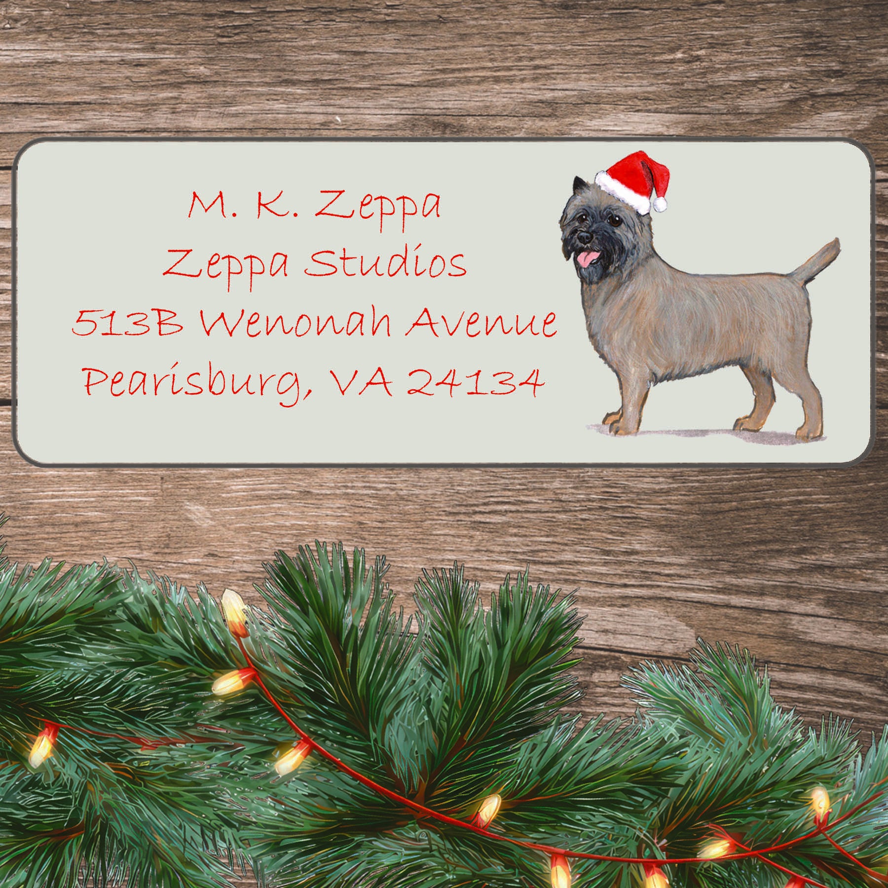 An illustration of a Cairn Terrier wearing a Santa Hat on an ivory return address label set off by a wood surface with holiday greenery background.