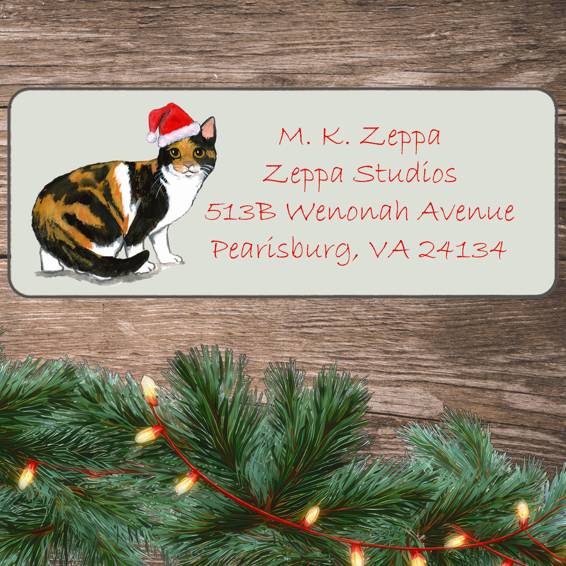 An illustration of a Calico Cat wearing a Santa Hat on an ivory return address label set off by a wood surface with holiday greenery background.