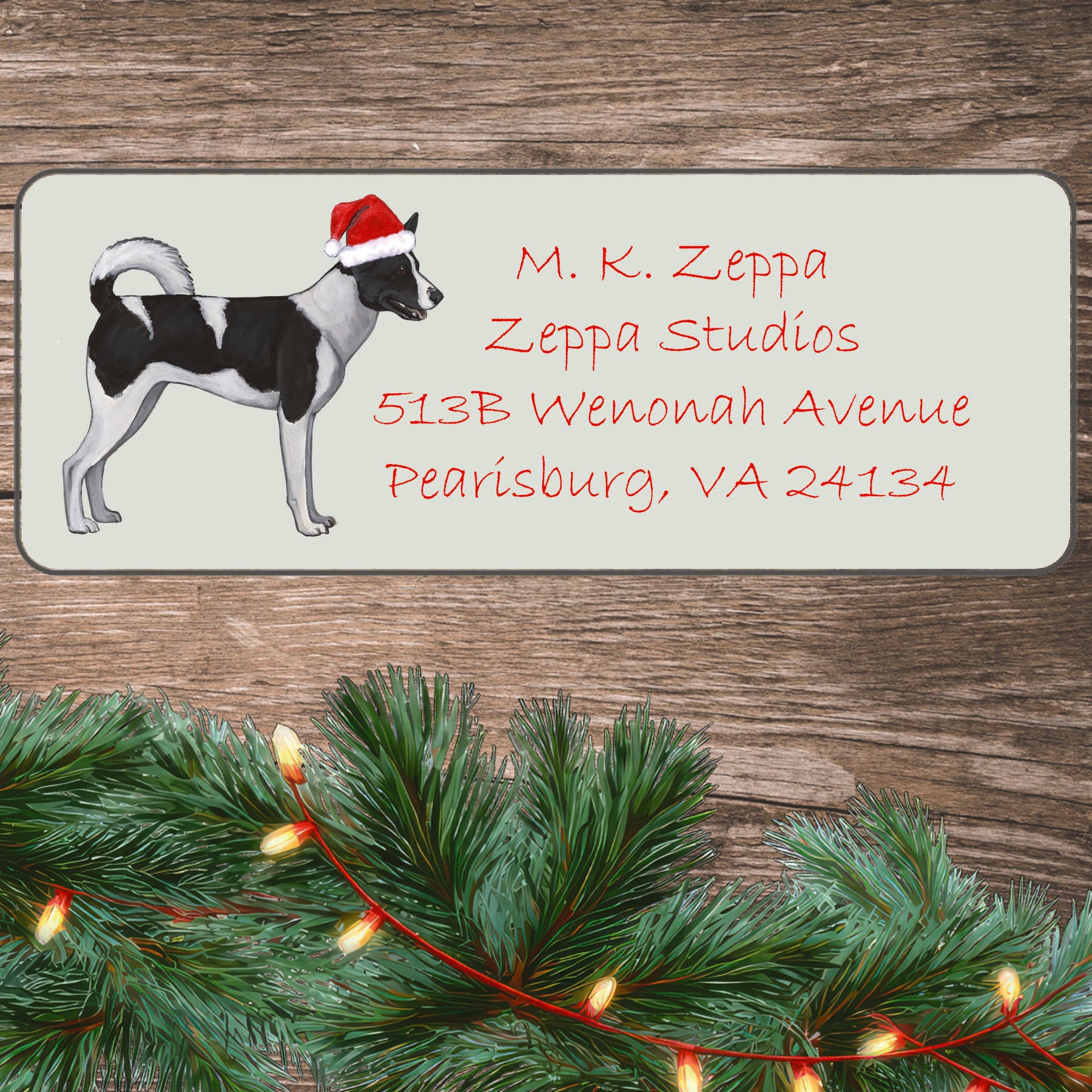 A Canaan Dog in a Santa Hat illustration on an ivory return address label set off by a wood surface with holiday greenery background.