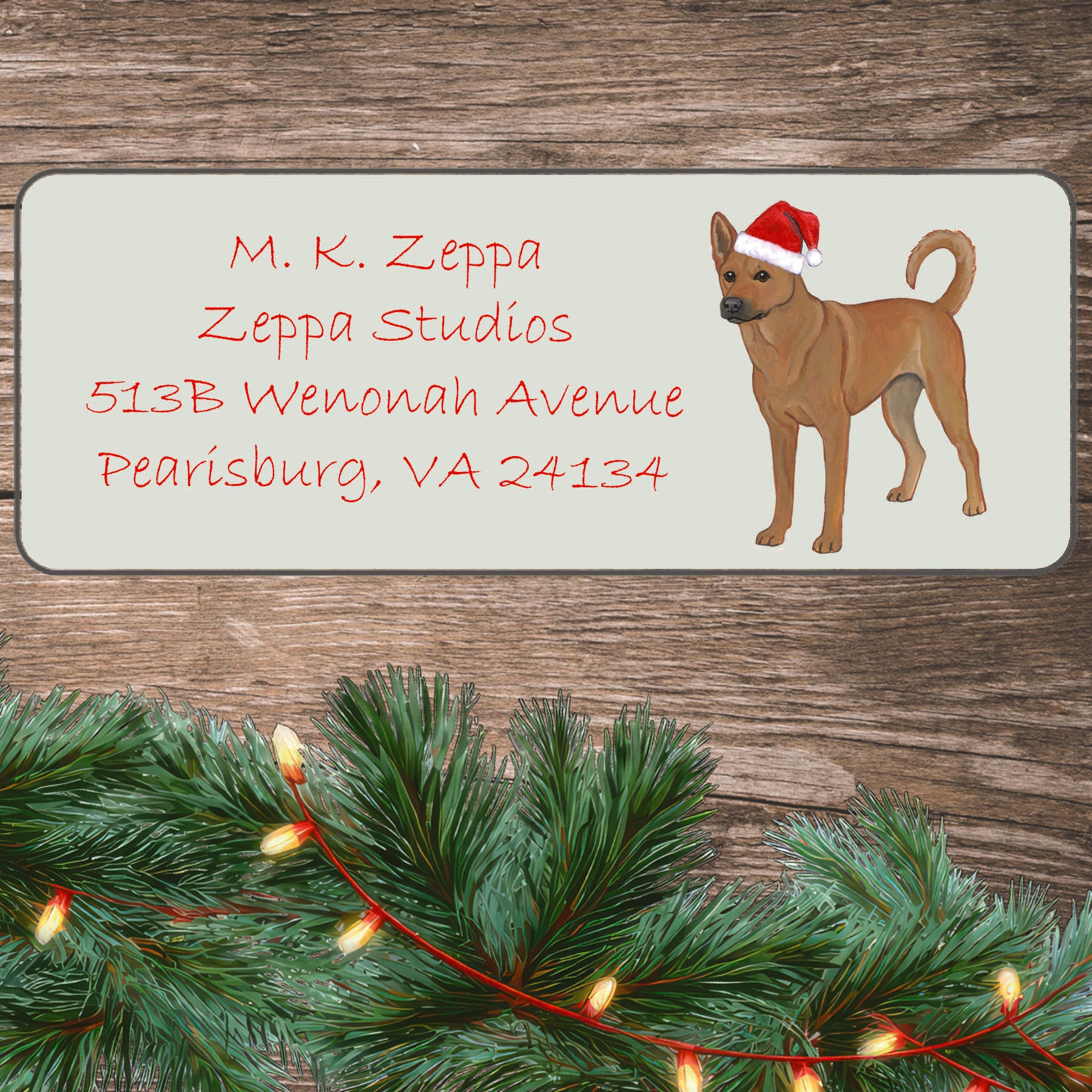 An illustration of a Carolina Dog wearing a Santa Hat on an ivory return address label set off by a wood surface with holiday greenery background.