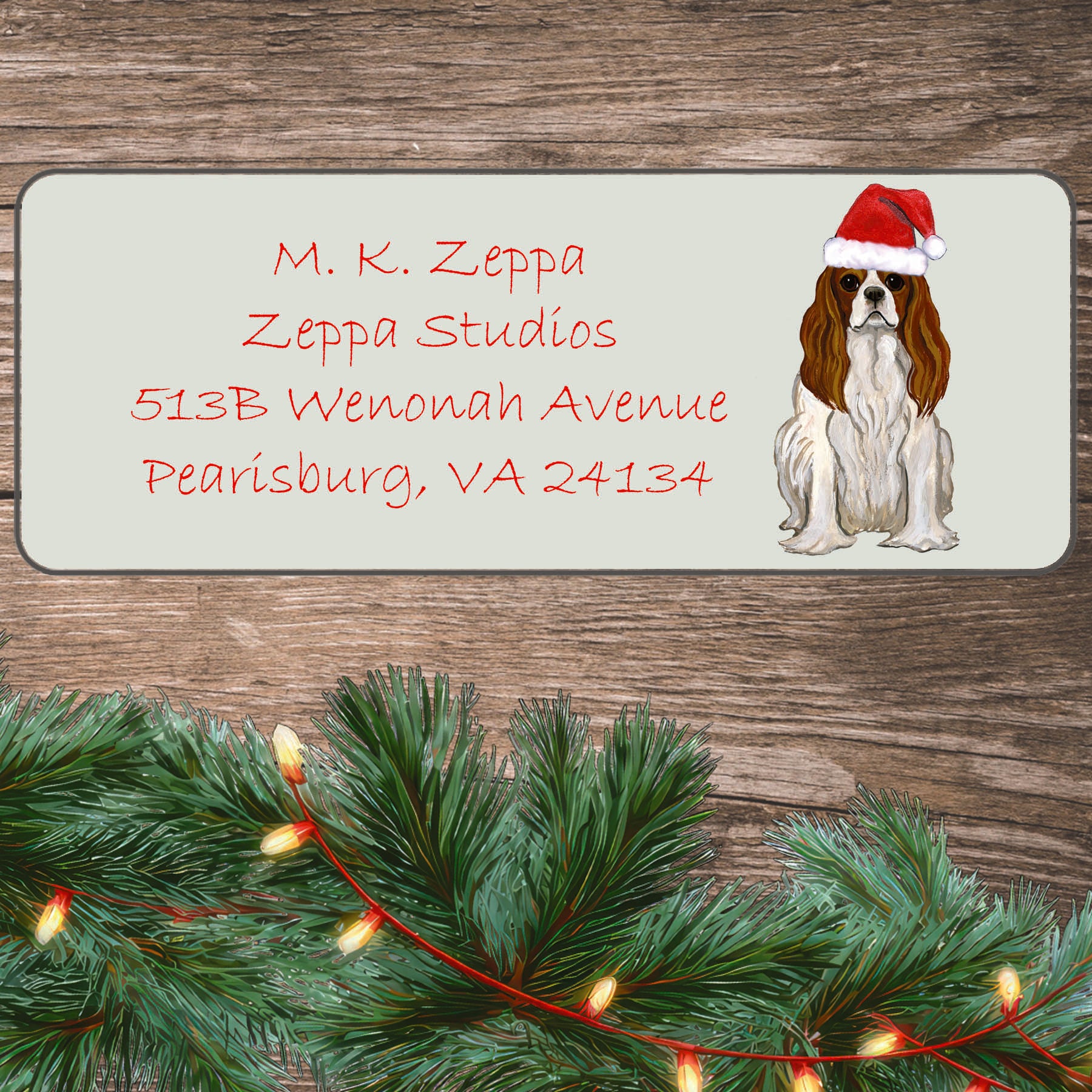 An illustration of a Blenheim Cavalier King Charles Spaniel wearing a Santa Hat on an ivory return address label set off by a wood surface with holiday greenery background.