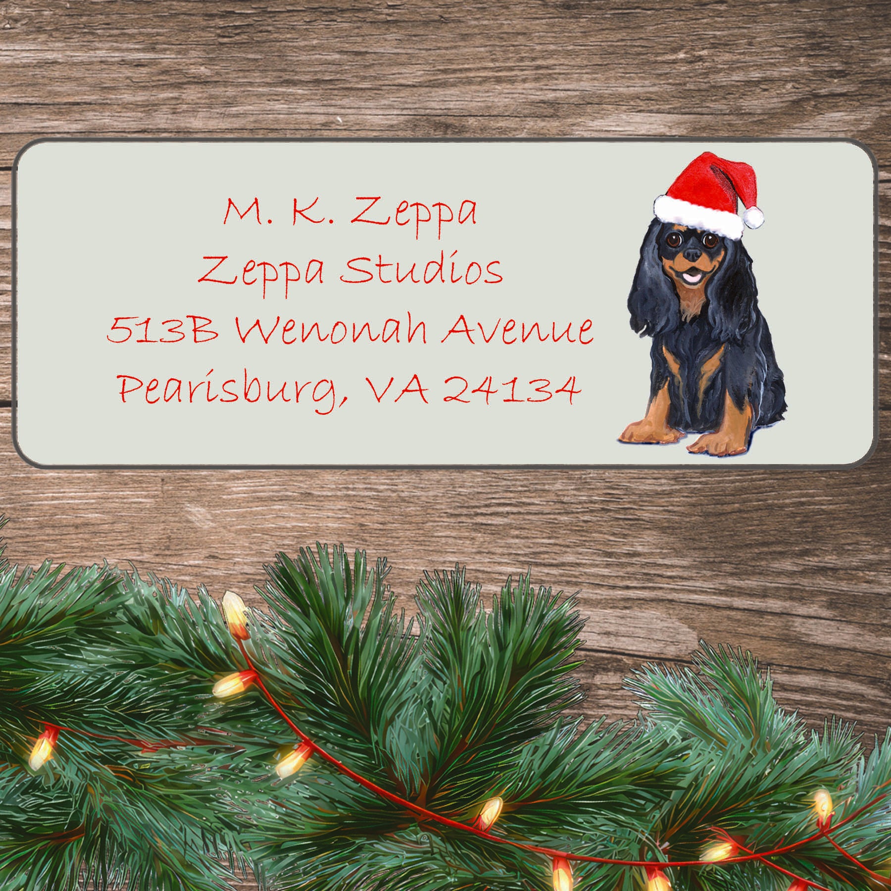 An illustration of a black and tan Cavalier King Charles Spaniel wearing a Santa Hat on an ivory return address label set off by a wood surface with holiday greenery background.
