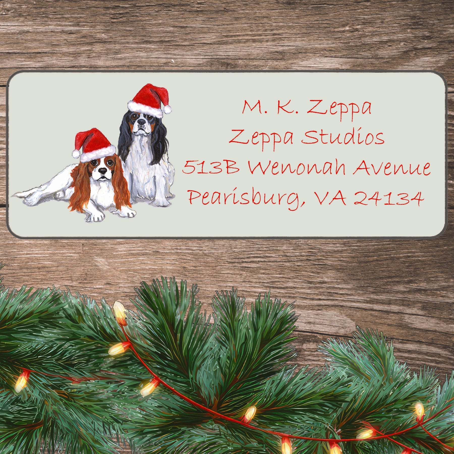 An illustration of a pair of Blenheim Cavalier King Charles Spaniels wearing Santa Hats on an ivory return address label set off by a wood surface with holiday greenery background.