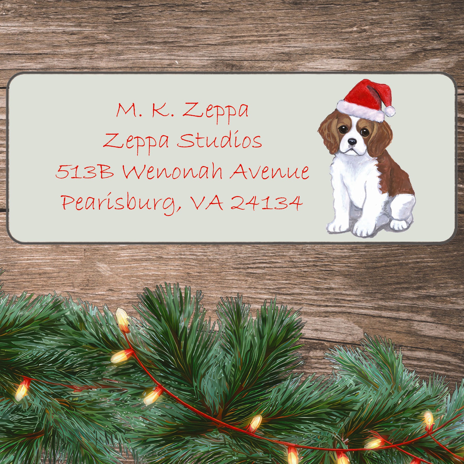 An illustration of a Cavalier King Charles Spaniel puppy wearing a Santa Hat on an ivory return address label set off by a wood surface with holiday greenery background.