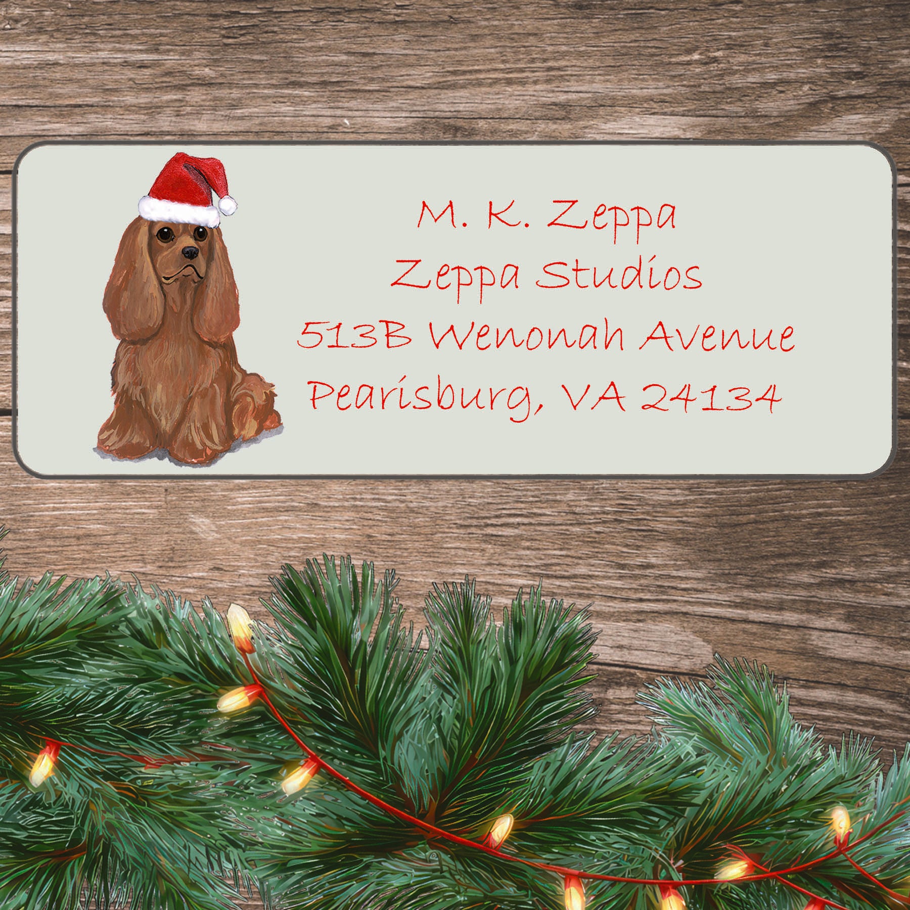 An illustration of a ruby Cavalier King Charles Spaniel wearing a Santa Hat on an ivory return address label set off by a wood surface with holiday greenery background.