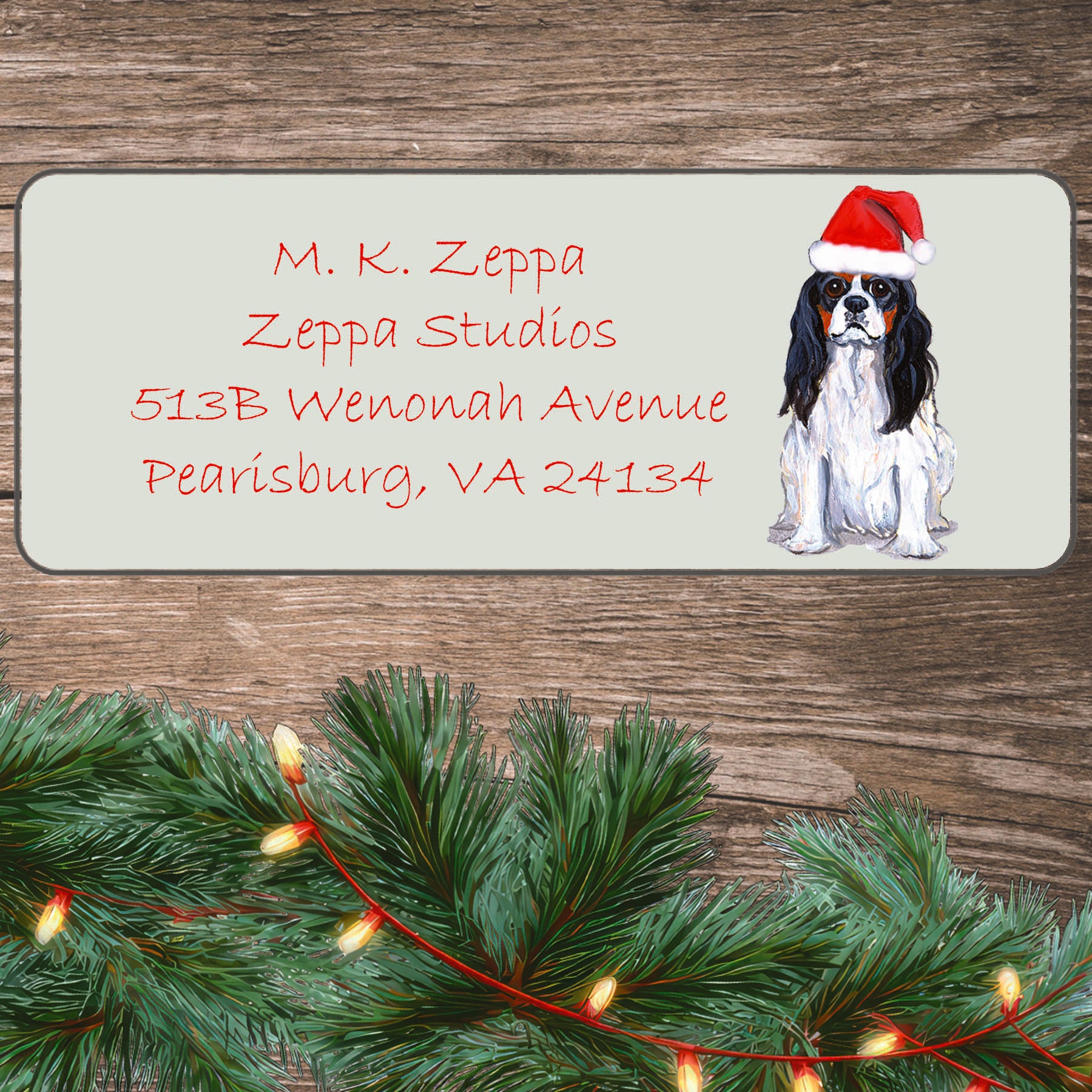 An illustration of a tri-color Cavalier King Charles Spaniel wearing a Santa Hat on an ivory return address label set off by a wood surface with holiday greenery background.