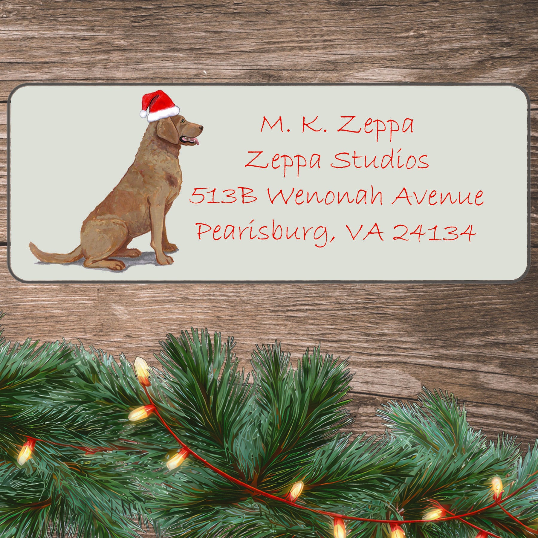 An illustration of a Chesapeake Bay  Retriever wearing a Santa Hat on an ivory return address label set off by a wood surface with holiday greenery background,