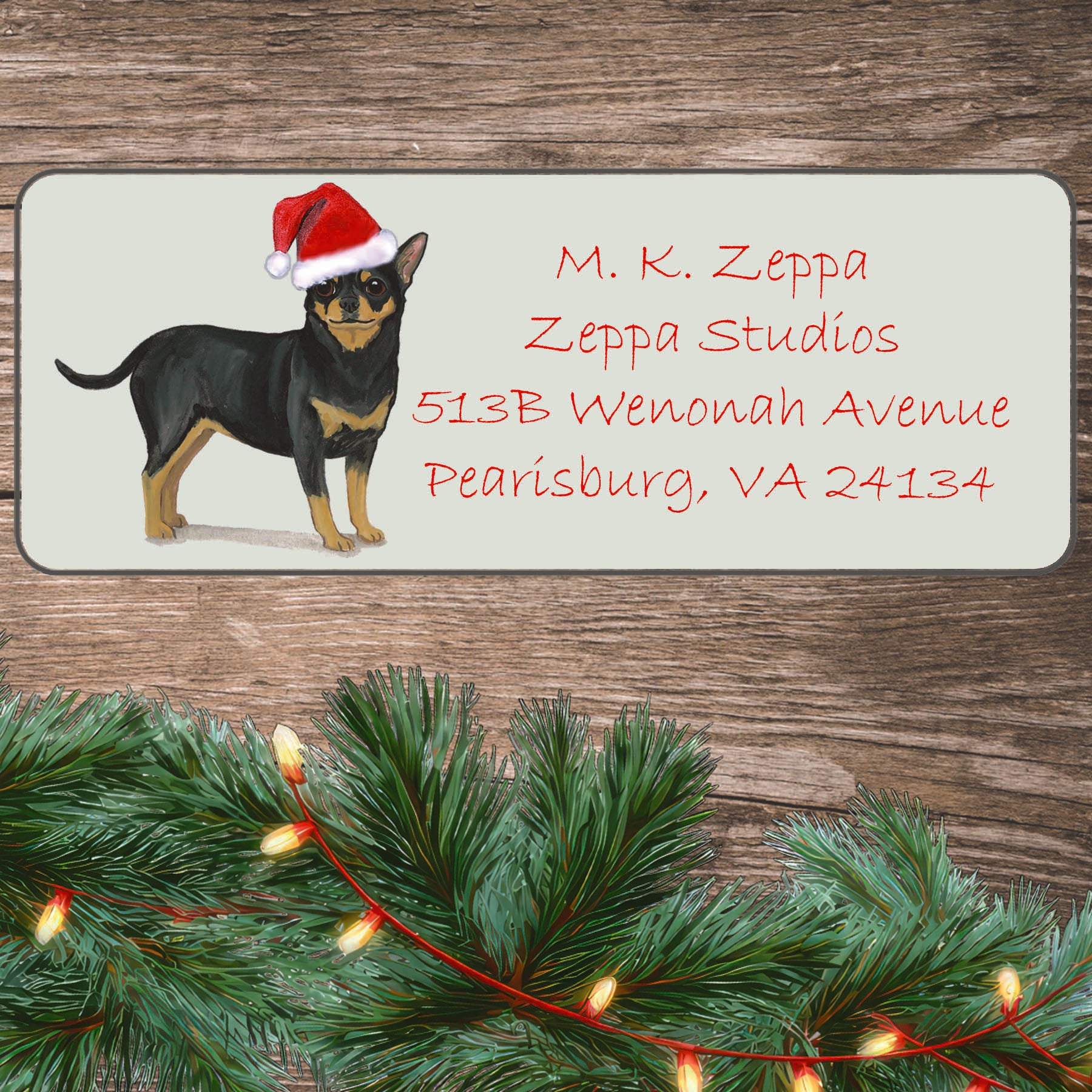 An illustration of a black and tan Chihuahua wearing a Santa Hat on an ivory return address label set off by a wood surface with holiday greenery background.