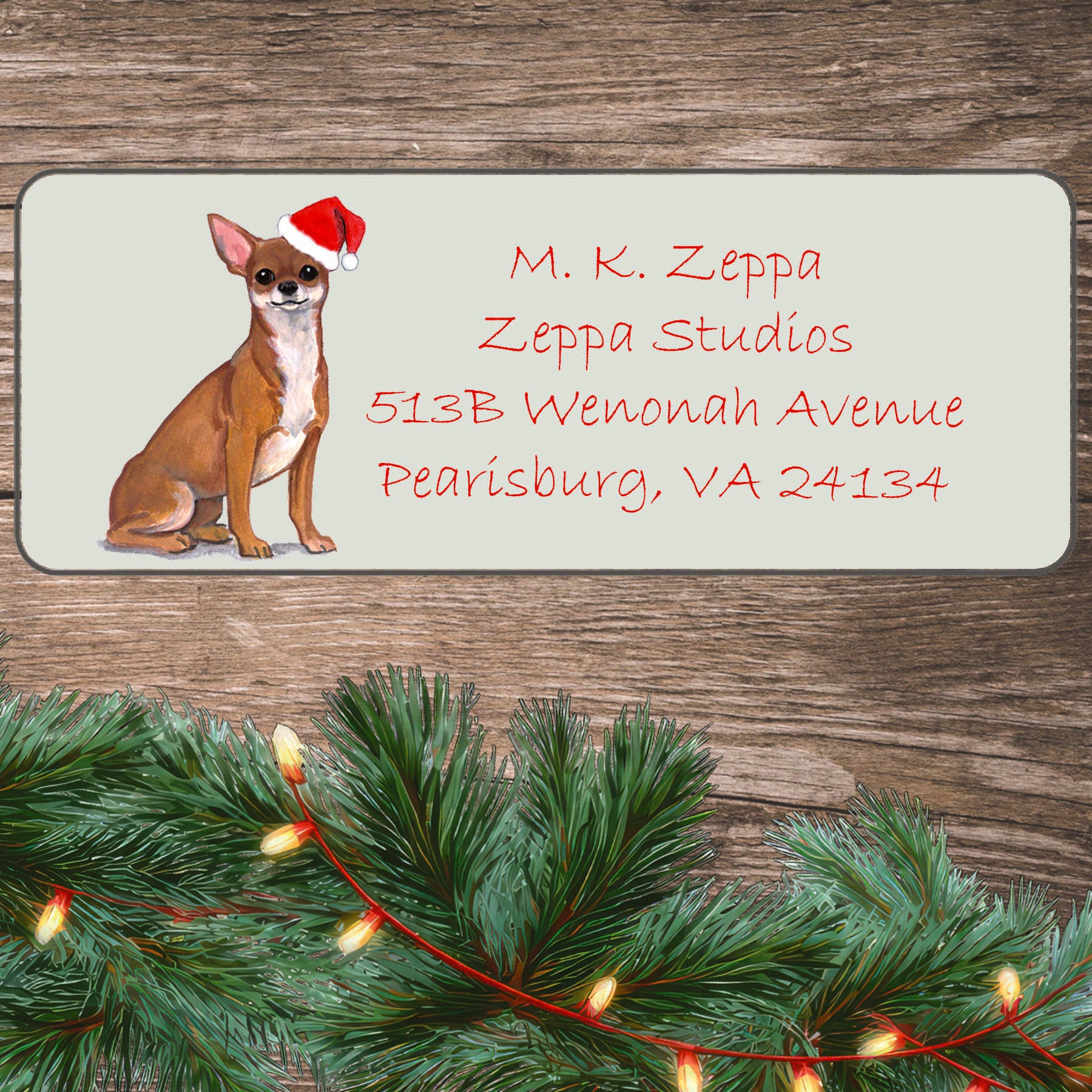 An illustration of a Chihuahua wearing a Santa Hat on an ivory return address label set off by a wood surface with holiday greenery background.
