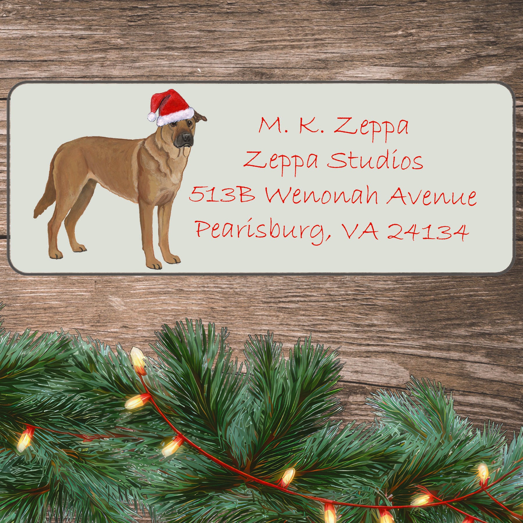 An illustration of a Chinook wearing a Santa Hat on an ivory return address label set off by a wood surface with holiday greenery background.