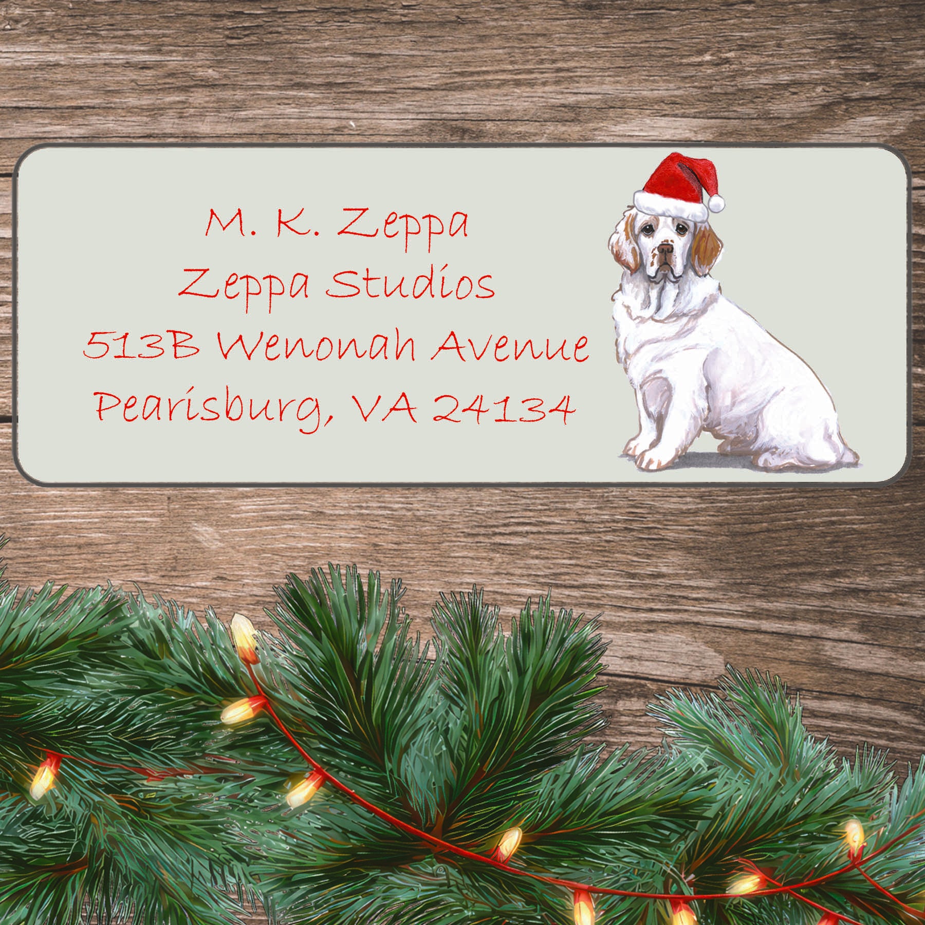 An illustration of a Clumber Spaniel wearing a Santa Hat on an ivory return address label set off by a wood surface with holiday greenery background.