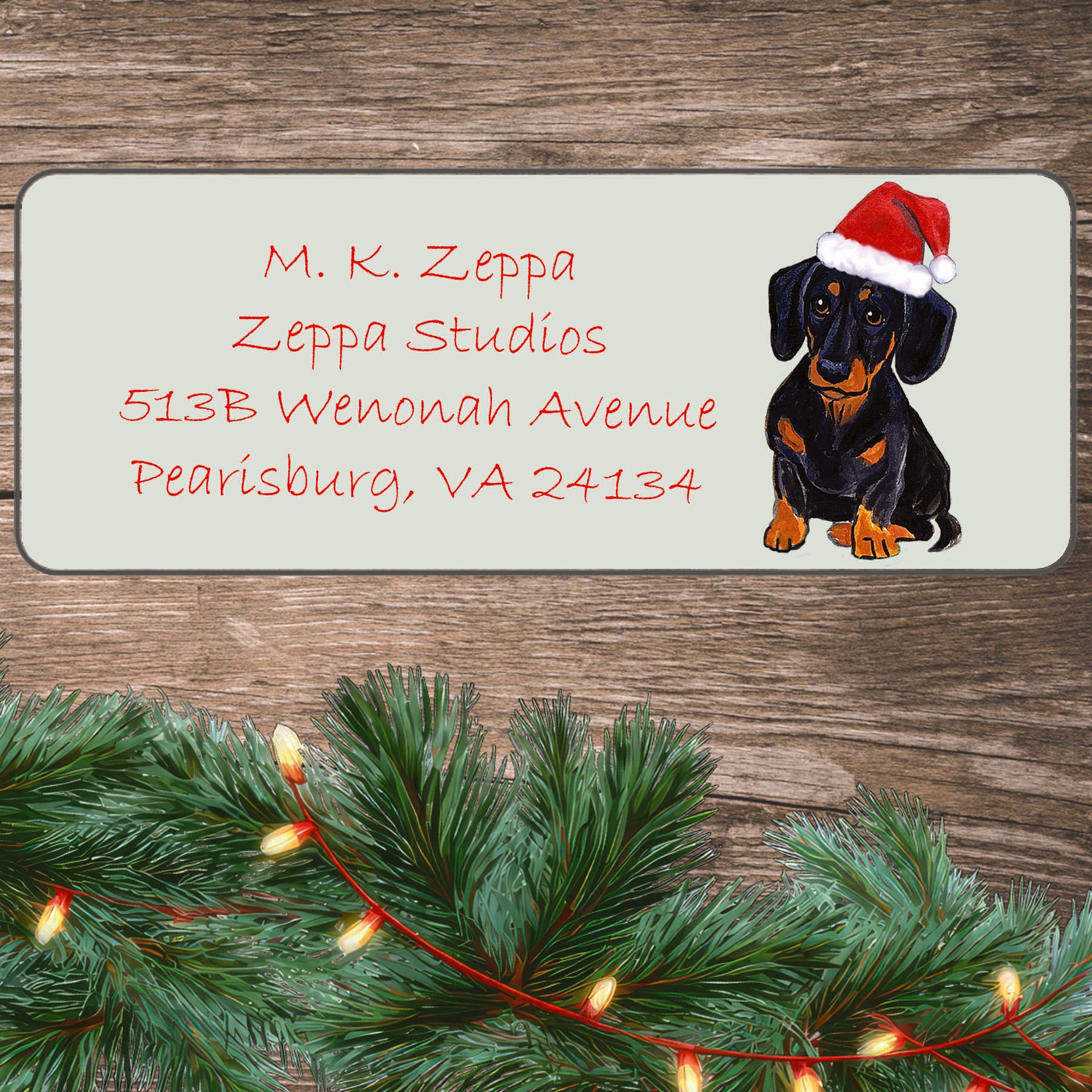 An illustration of a black and tan Dachshund wearing a Santa Hat on an ivory return address label set off by a wood surface with holiday greenery background.