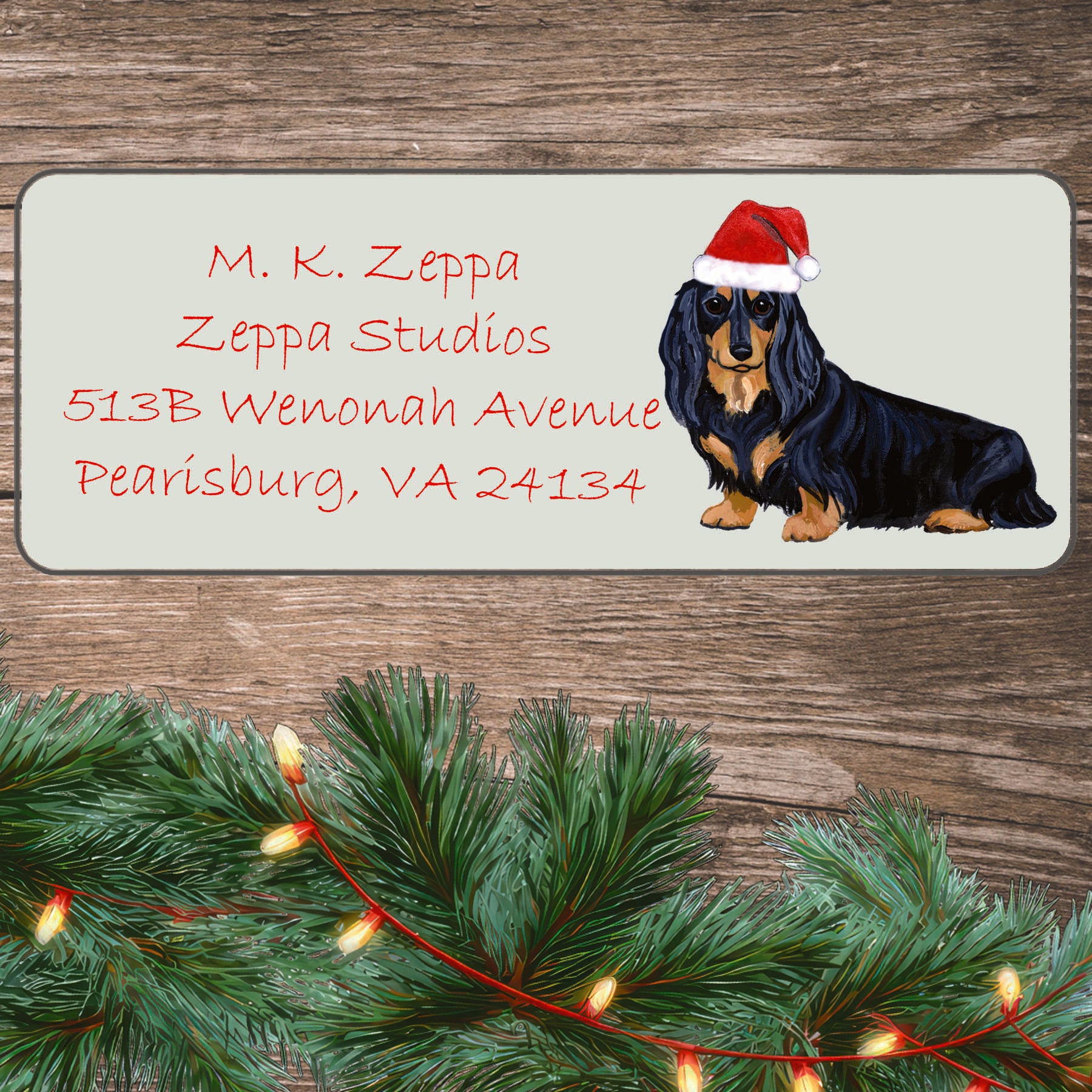An illustration of a longhaired black and tan Dachshund wearing a Santa Hat on an ivory return address label set off by a wood surface with holiday greenery background.