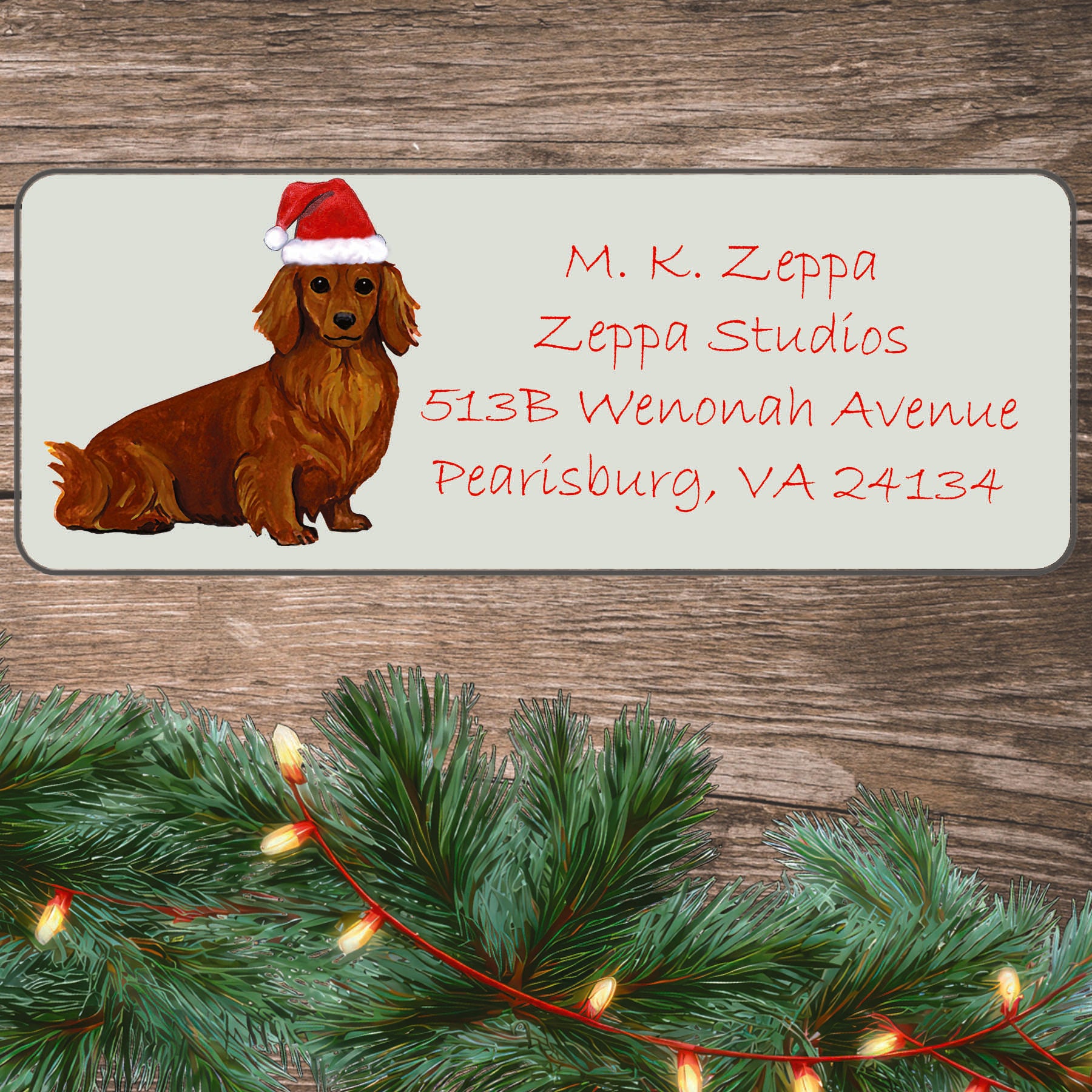 An illustration of a red long-haired Dachshund wearing a Santa Hat on an ivory return address label set off by a wood surface with holiday greenery background.