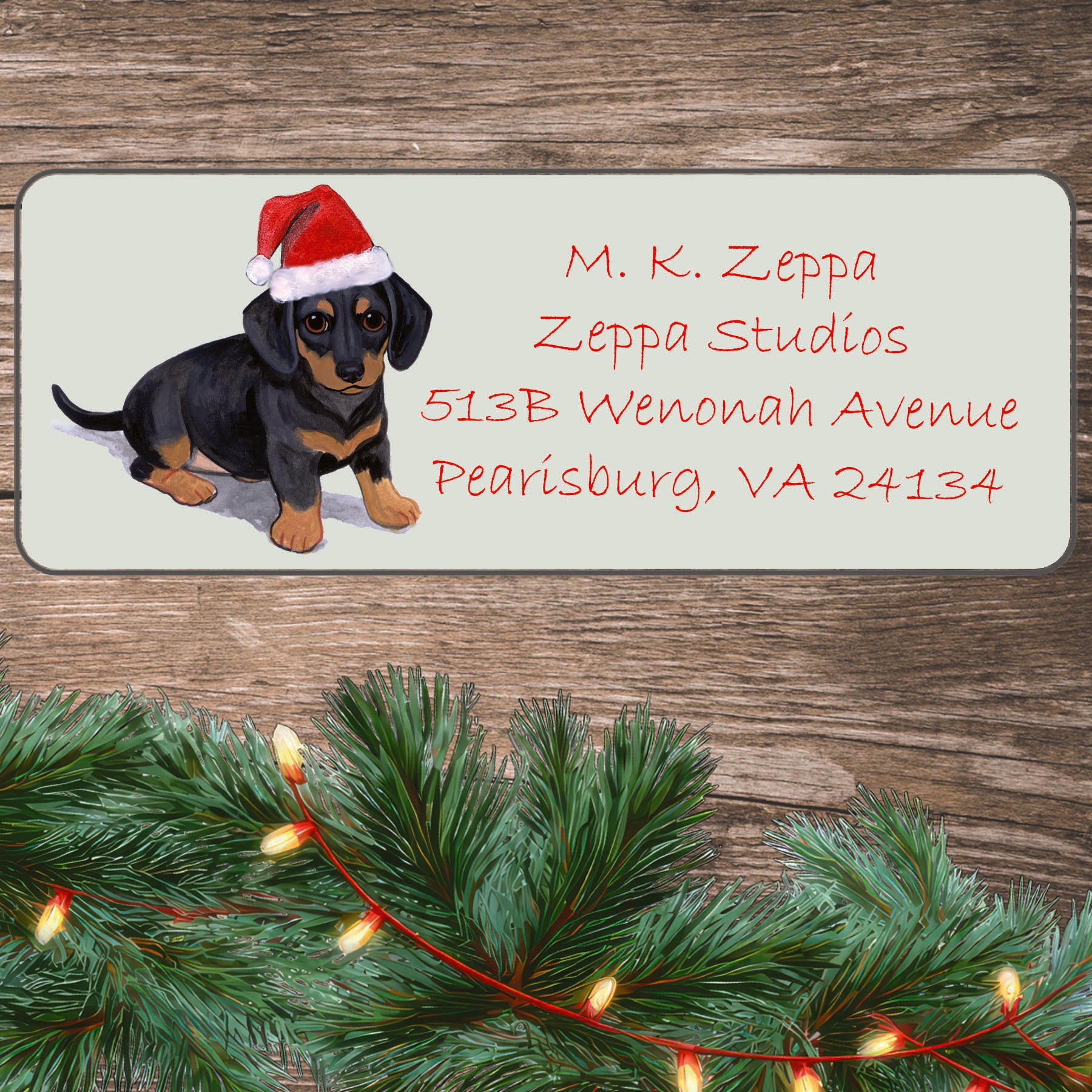An illustration of a Dachshund puppy wearing a Santa Hat on an ivory return address label set off by a wood surface with holiday greenery background.