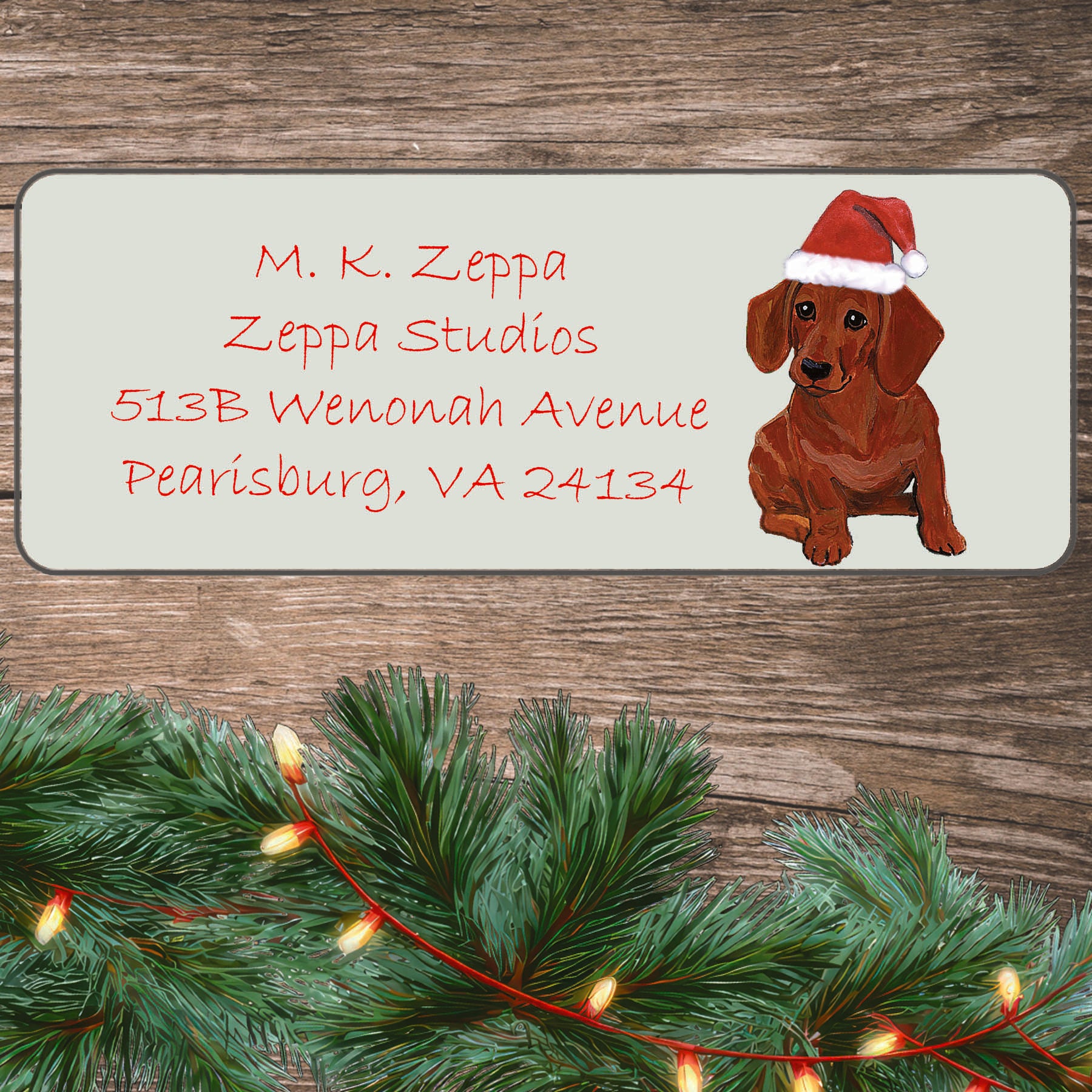 An illustration of a red Dachshund wearing a Santa Hat on an ivory return address label set off by a wood surface with holiday greenery background.