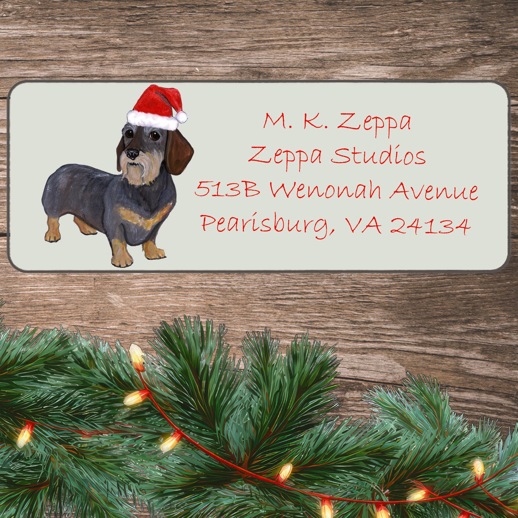 An illustration of a Wirehaired Dachshund wearing a Santa Hat on an ivory return address label set off by a wood surface with holiday greenery background.