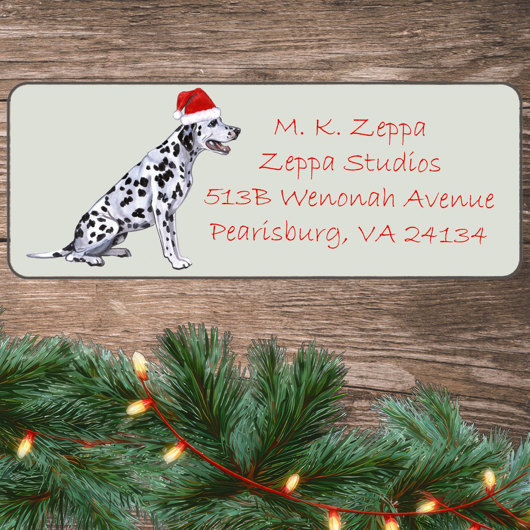 An illustration of a Dalmatian wearing a Santa Hat on an ivory return address label set off by a wood surface with holiday greenery background.