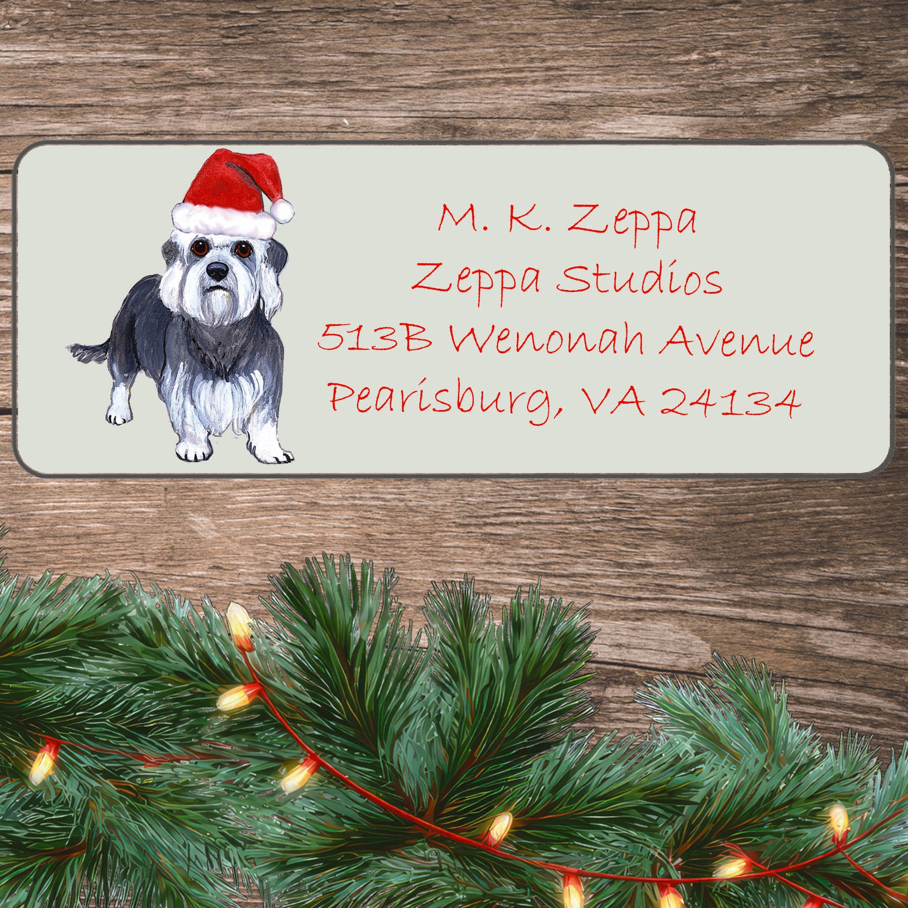 An illustration of a Dandi Dinmont Terrier wearing a Santa Hat on an ivory return address label set off by a wood surface with holiday greenery background.