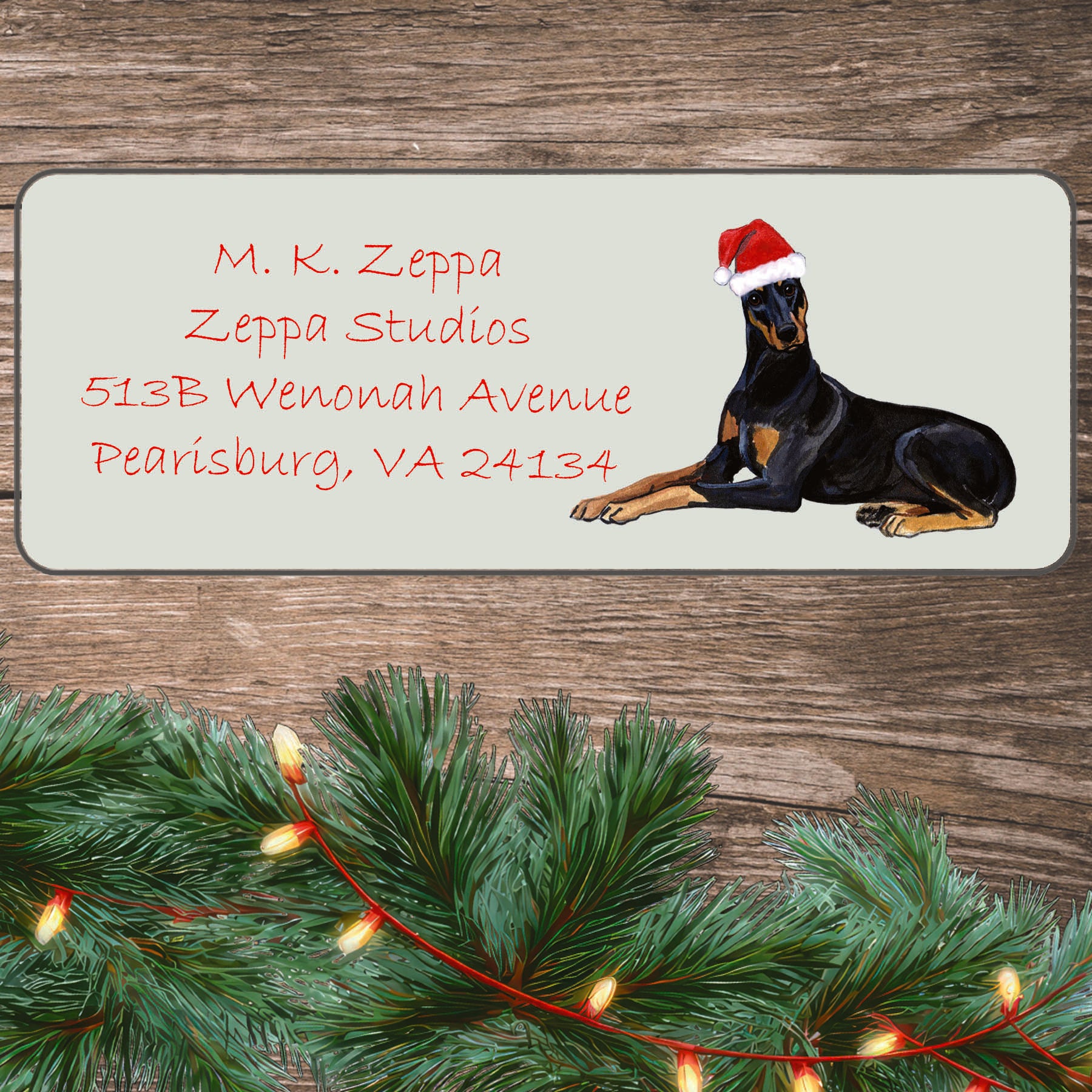 An illustration of a Doberman Pinscher wearing a Santa Hat on an ivory return address label set off by a wood surface with holiday greenery background.