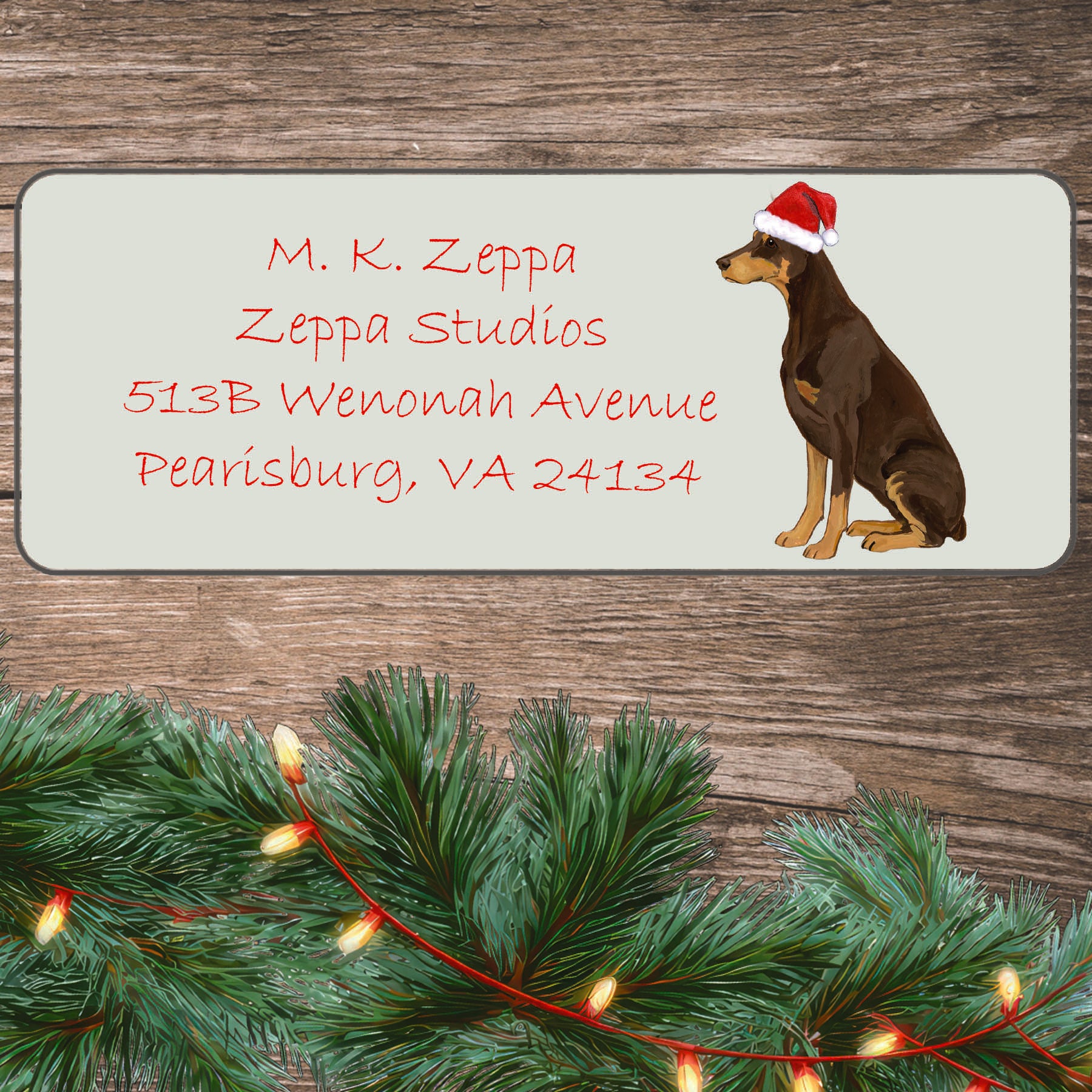 An illustration of a red Doberman wearing a Santa Hat on an ivory return address label set off by a wood surface with holiday greenery background.