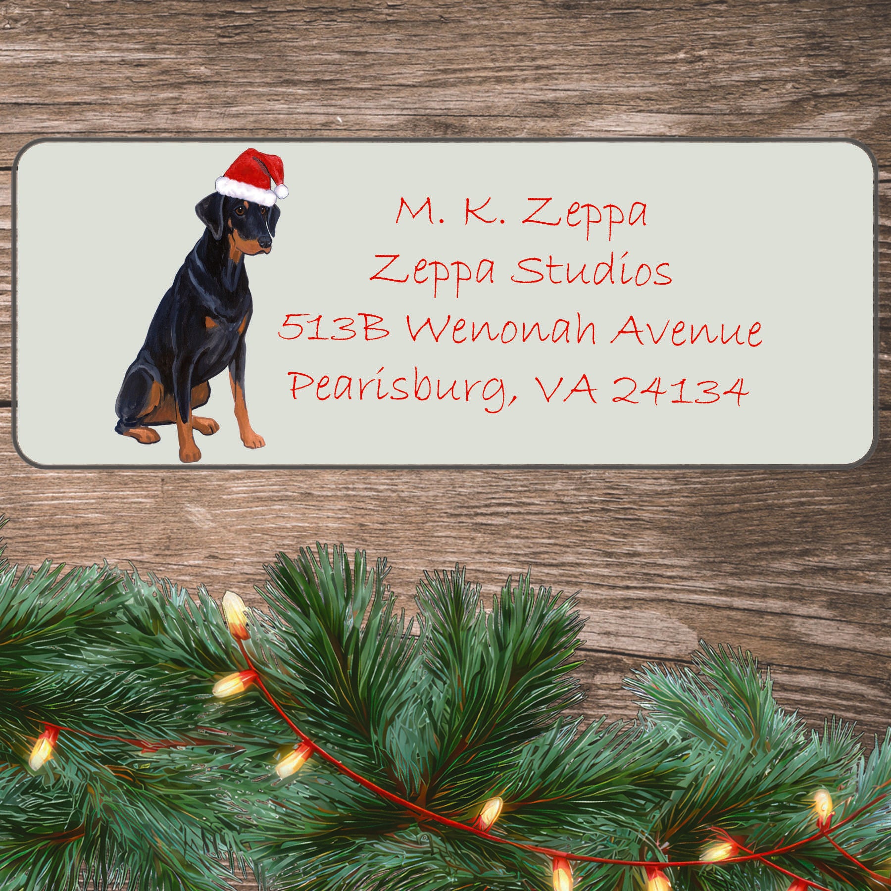 An illustration of a Doberman with natural ears wearing a Santa Hat on an ivory return address label set off by a wood surface with holiday greenery background.