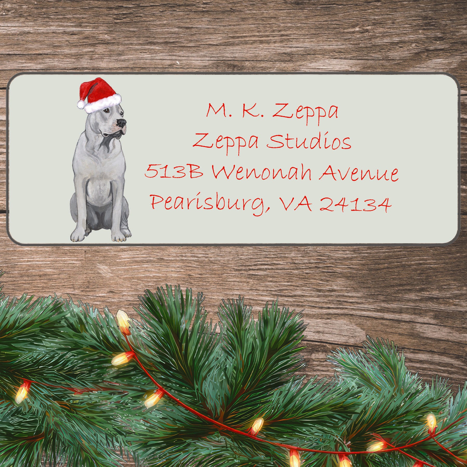 A Dogo Argentino in a Santa Hat illustration on an ivory return address label set off by a wood surface with holiday greenery background.