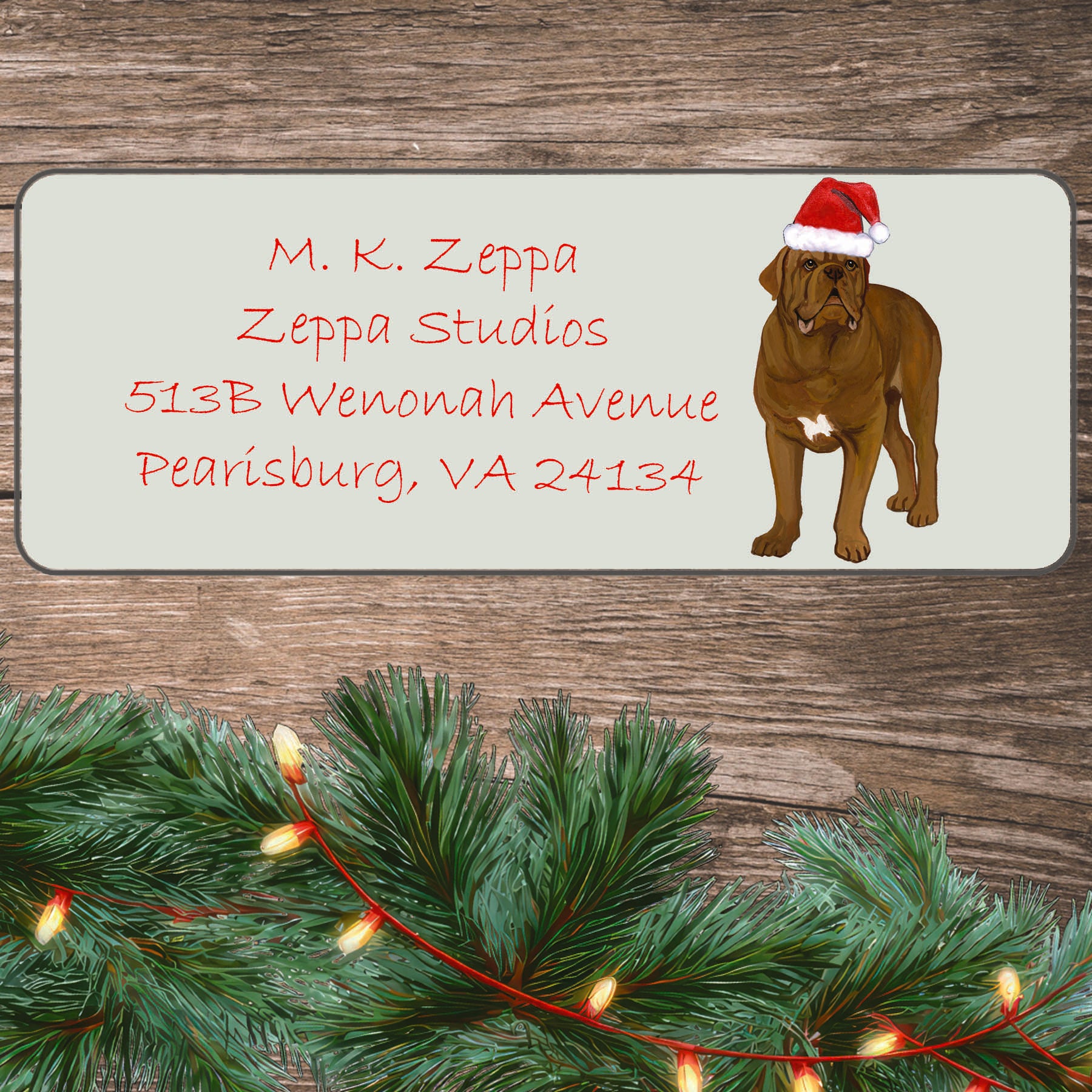 An illustration of a Dogue de Bordeaux wearing a Santa Hat on an ivory return address label set off by a wood surface with holiday greenery background. 