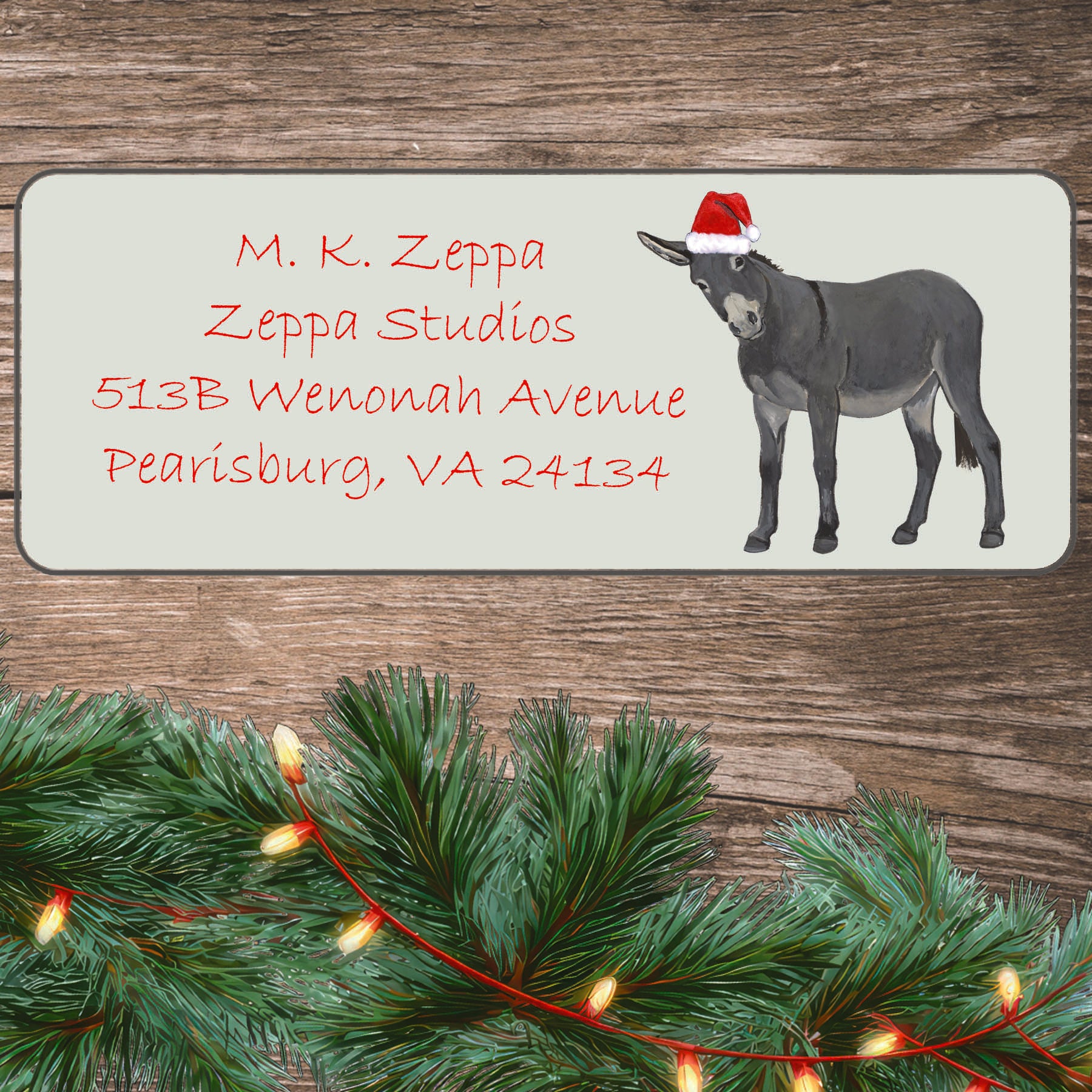 An illustration of a Donkey wearing a Santa Hat on an ivory return address label set off by a wood surface with holiday greenery background.
