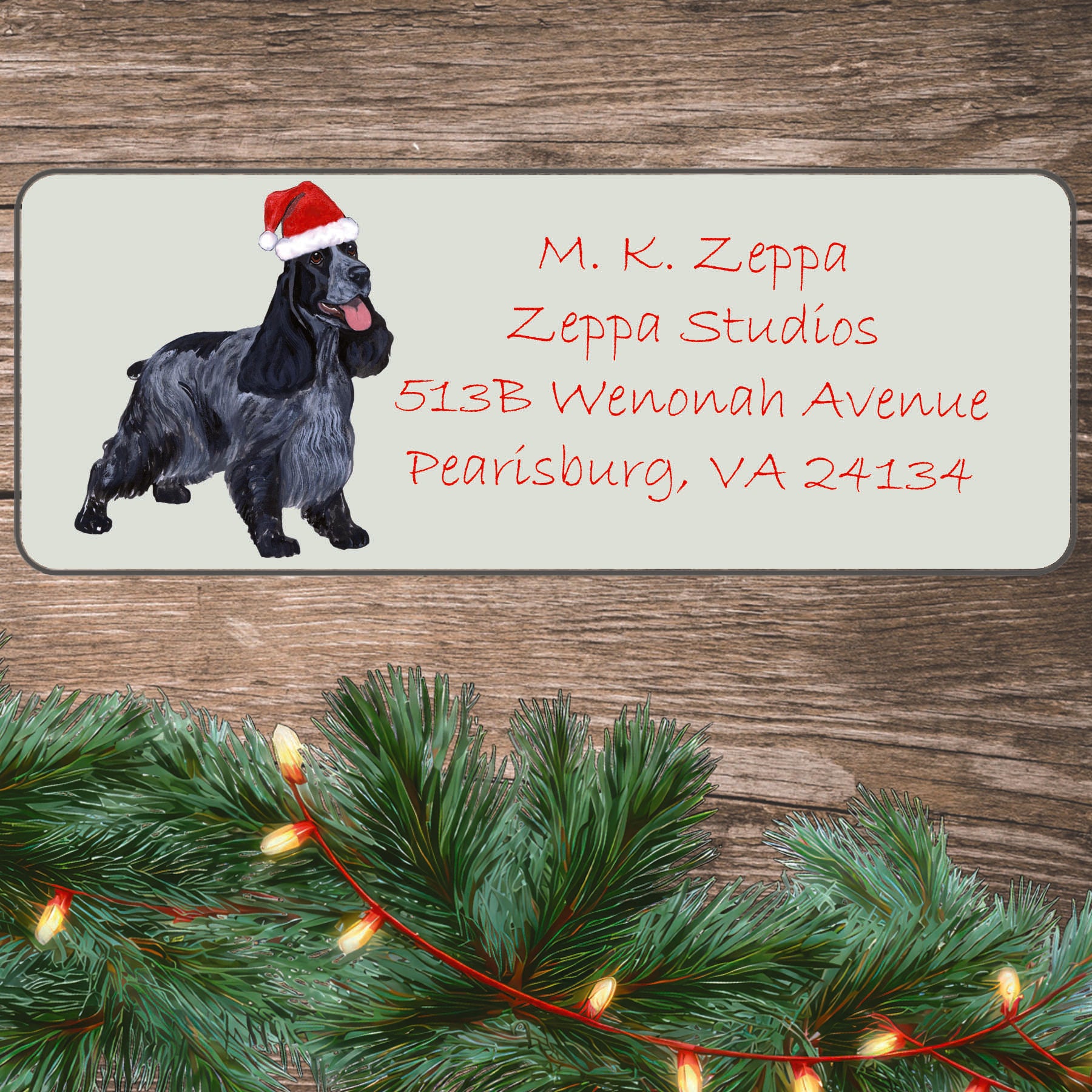 An illustration of an English Cocker Spaniel wearing a Santa Hat on an ivory return address label set off by a wood surface with holiday greenery background.