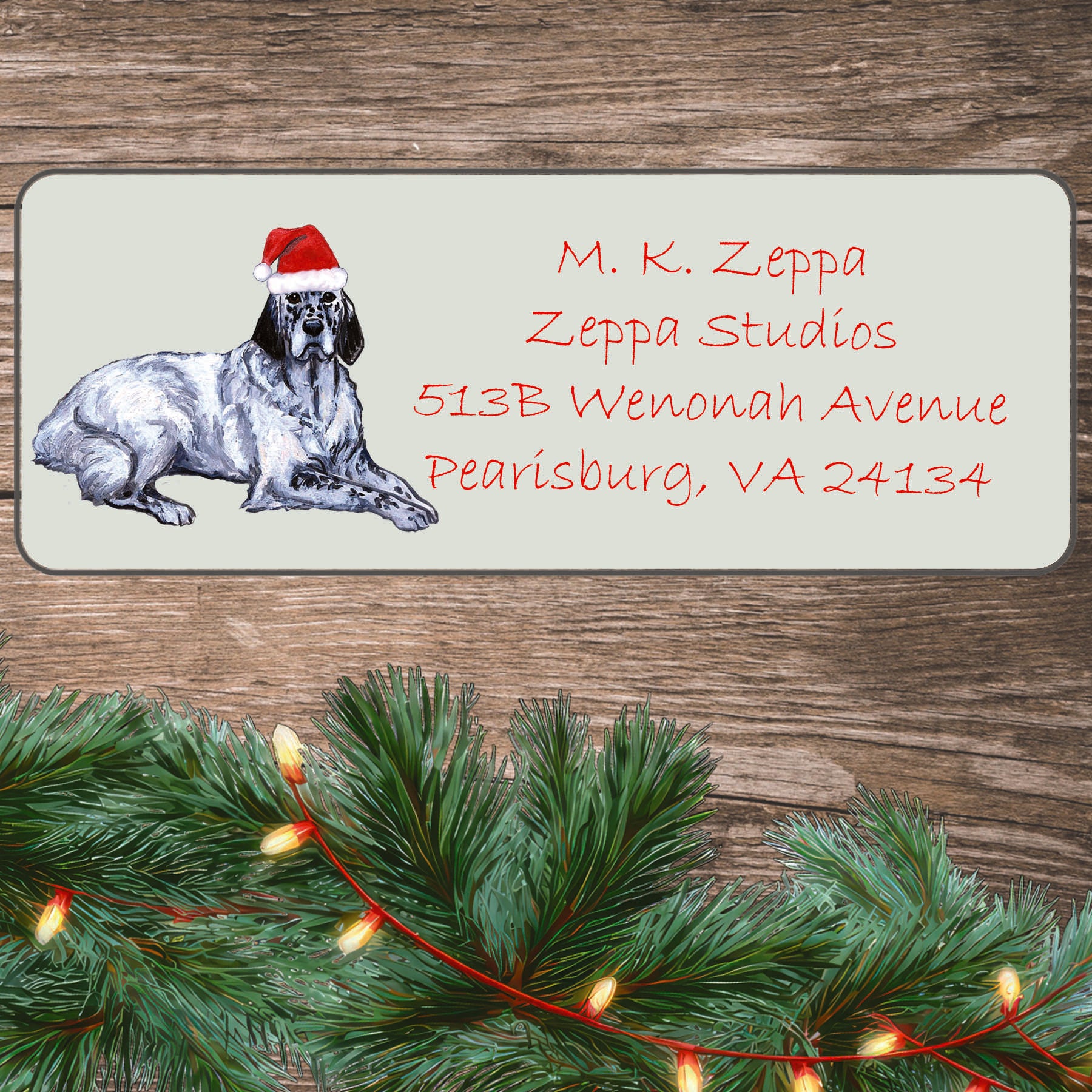 An illustration of a blue belton English Setter  wearing a Santa Hat on an ivory return address label set off by a wood surface with holiday greenery background.