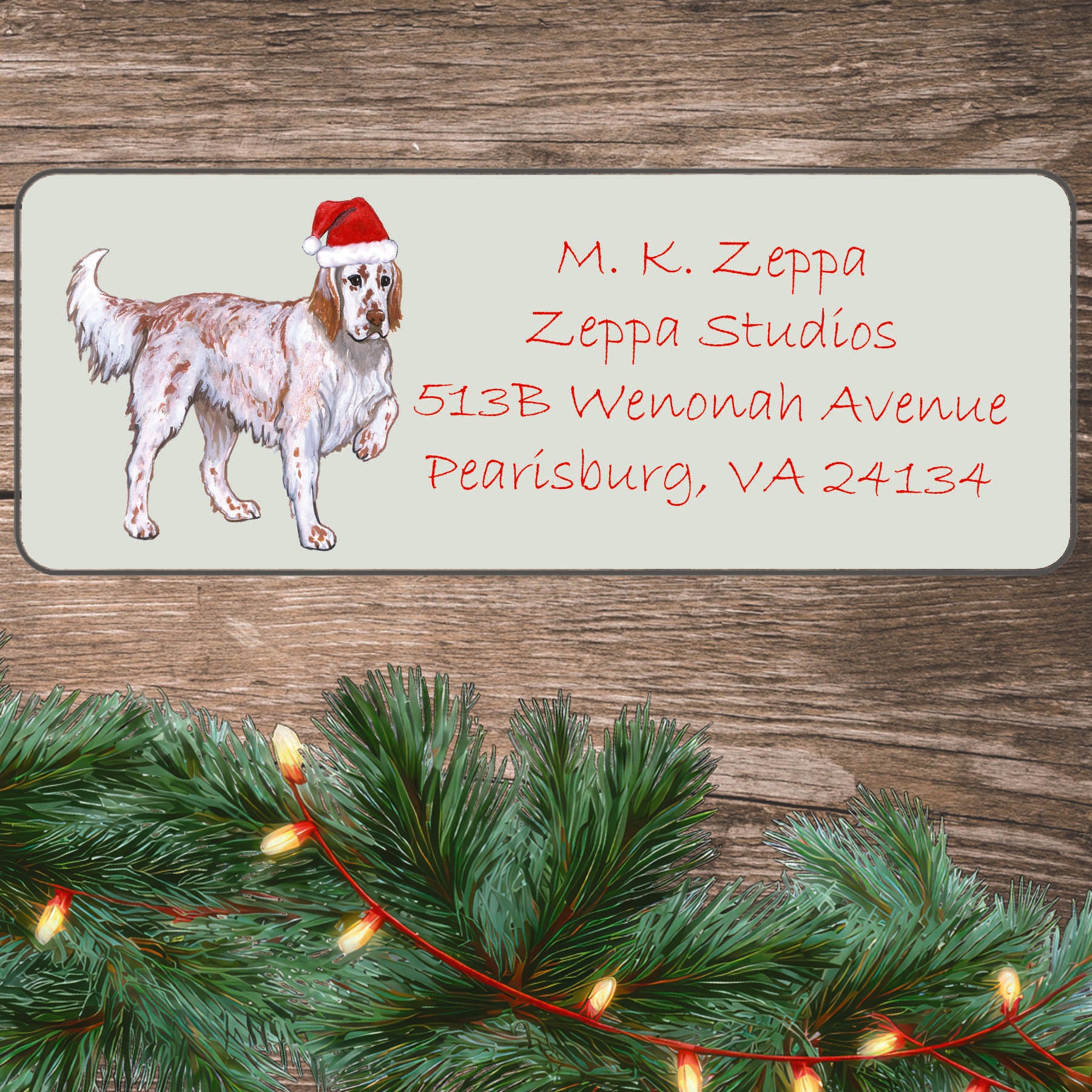 An illustration of a red belton English Setter wearing a Santa Hat on an ivory return address label set off by a wood surface with holiday greenery background.